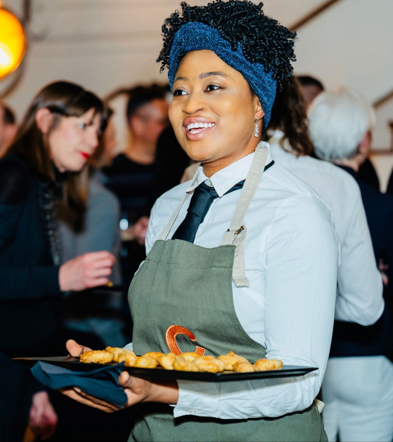 A smiling waitress in a white shirt, black tie, and green apron holds a tray of appetizers, with blurred people in the background. 