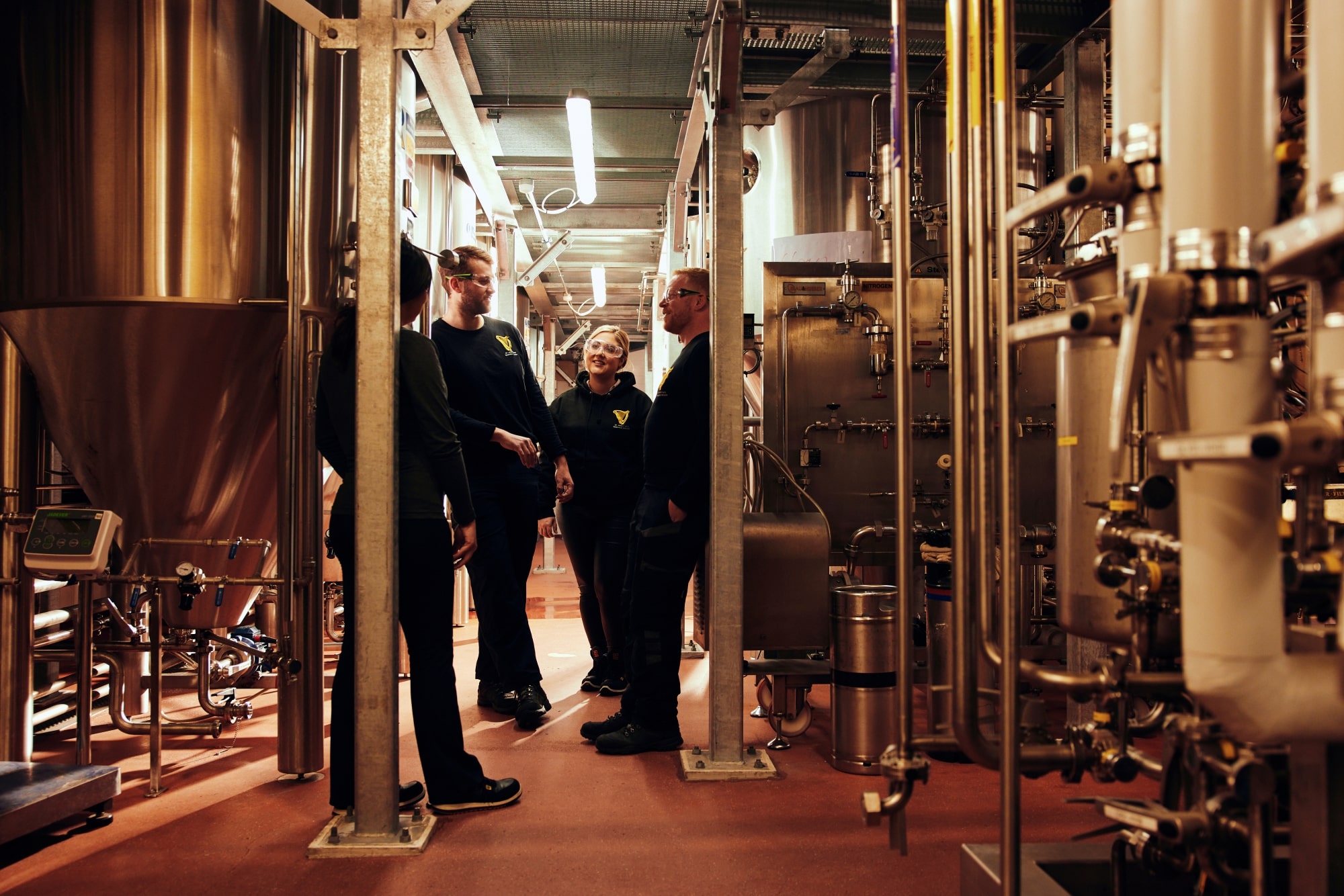 A group of four people, two men and two women, stand chatting in a brewery.