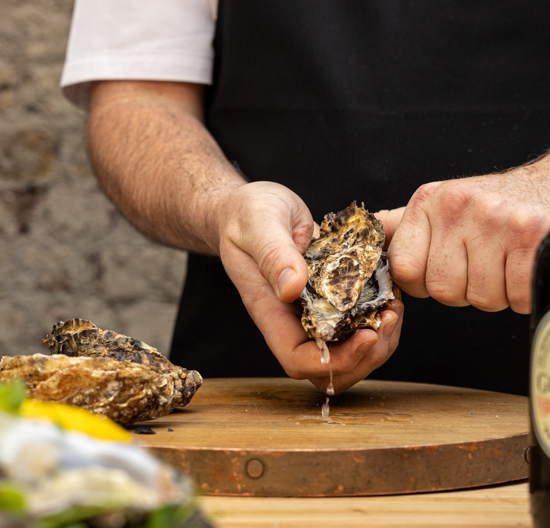 Hands shucking an oyster on a wooden cutting board, with other oysters and a bottle partially visible. 