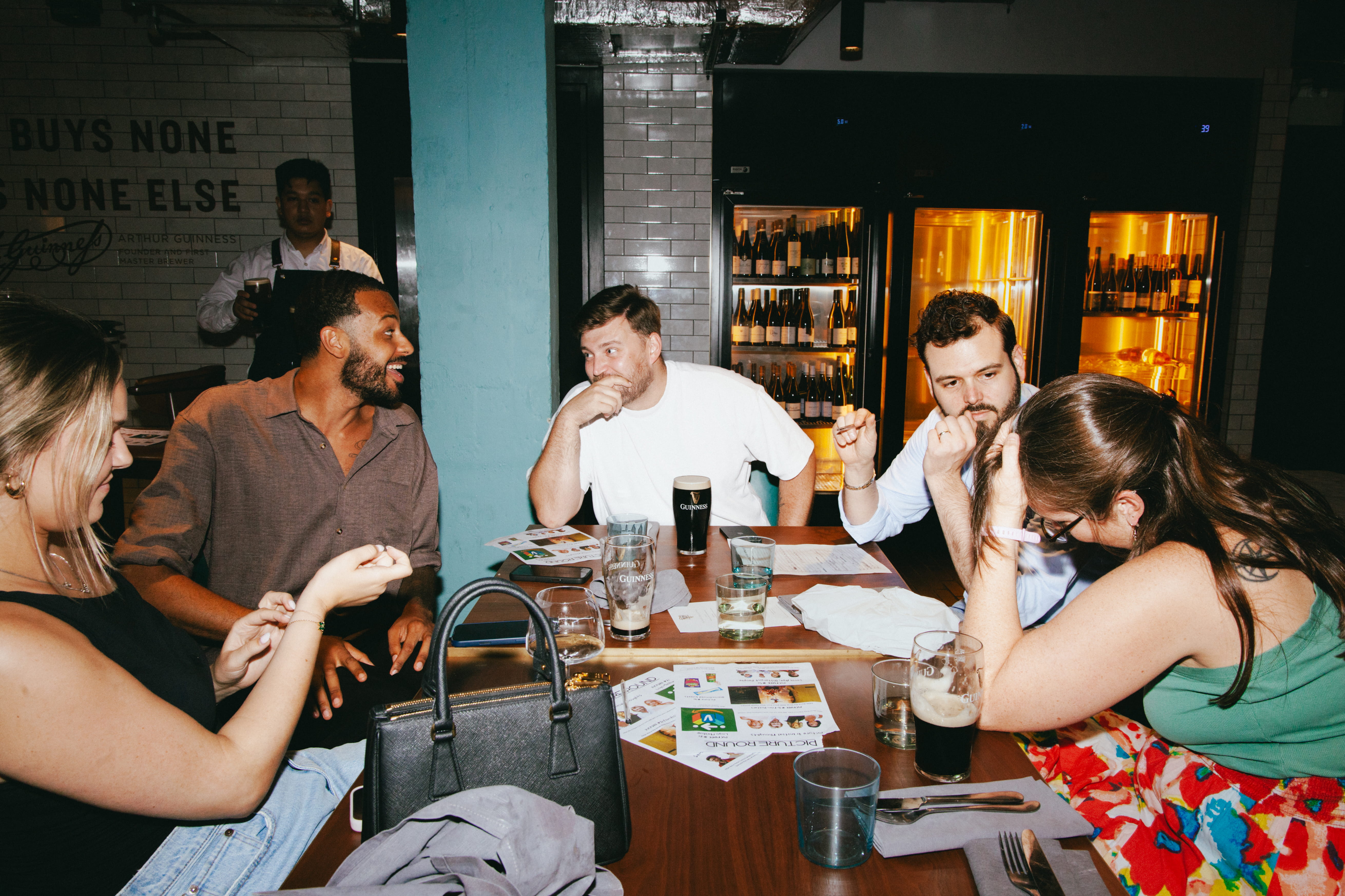 A group of people sat around a brown wooden table in a pub talking, with pints of Guinness and quiz sheets on the table