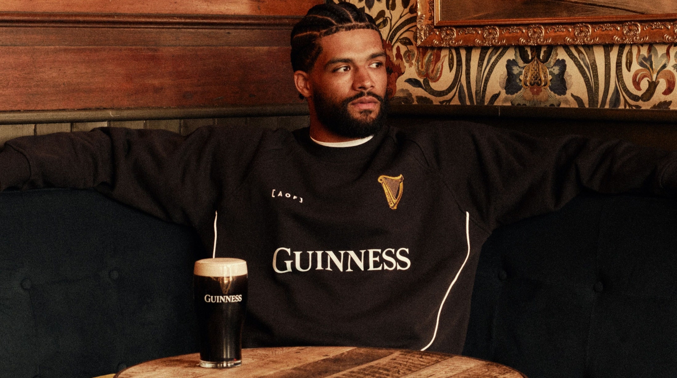A man with a beard and braided hair sits on a dark blue sofa, wearing a black Guinness sweatshirt, with a glass of Guinness beer on a wooden table. 