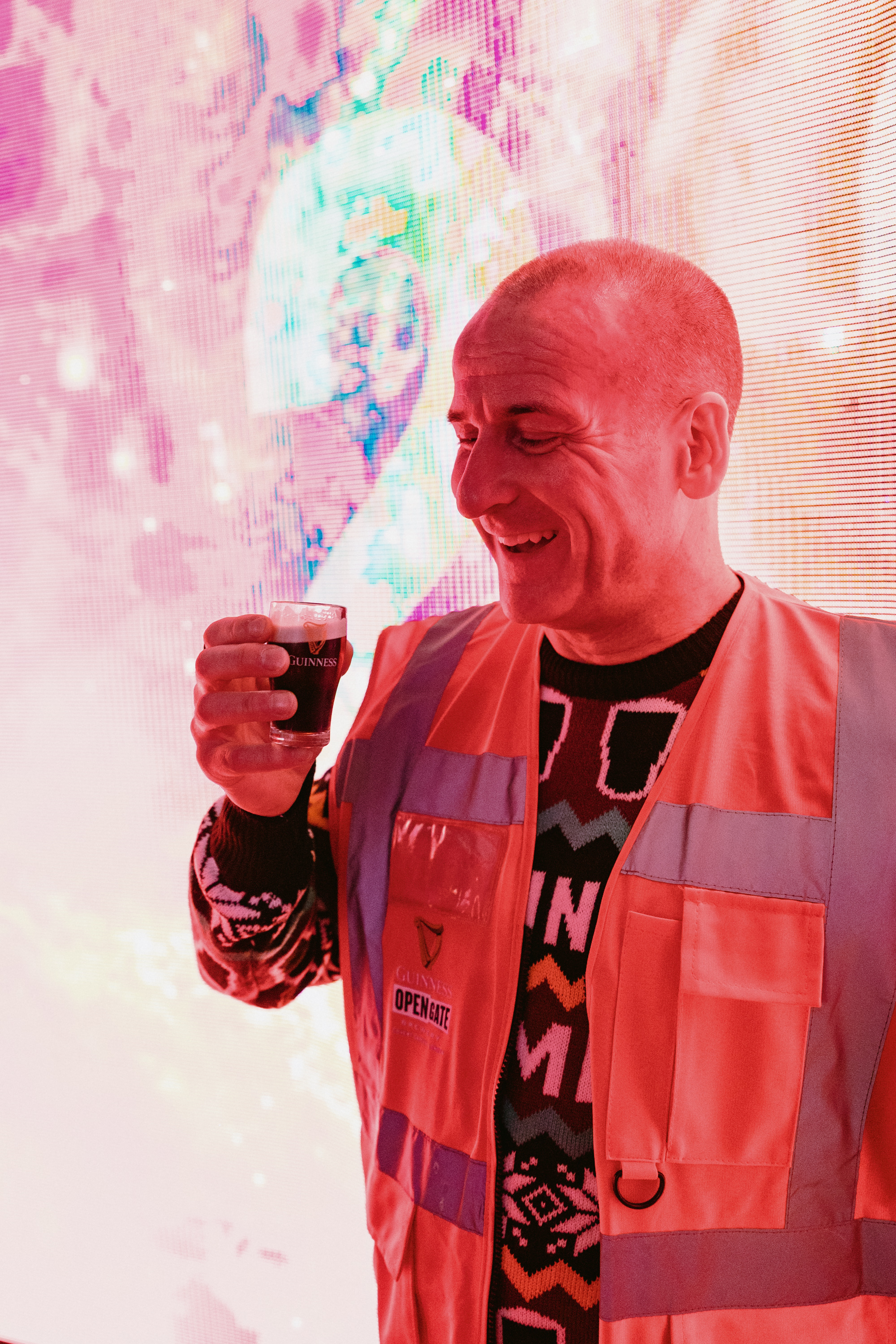 A man standing in a bright room wearing a Christmas jumper and a high-vis jacket holding a small Guinness shop glass
