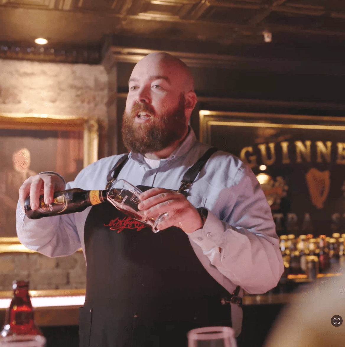 A bearded man in a light blue shirt and black apron pours a drink from a bottle into a glass behind a bar, with a "GUINNESS" sign in the background. 