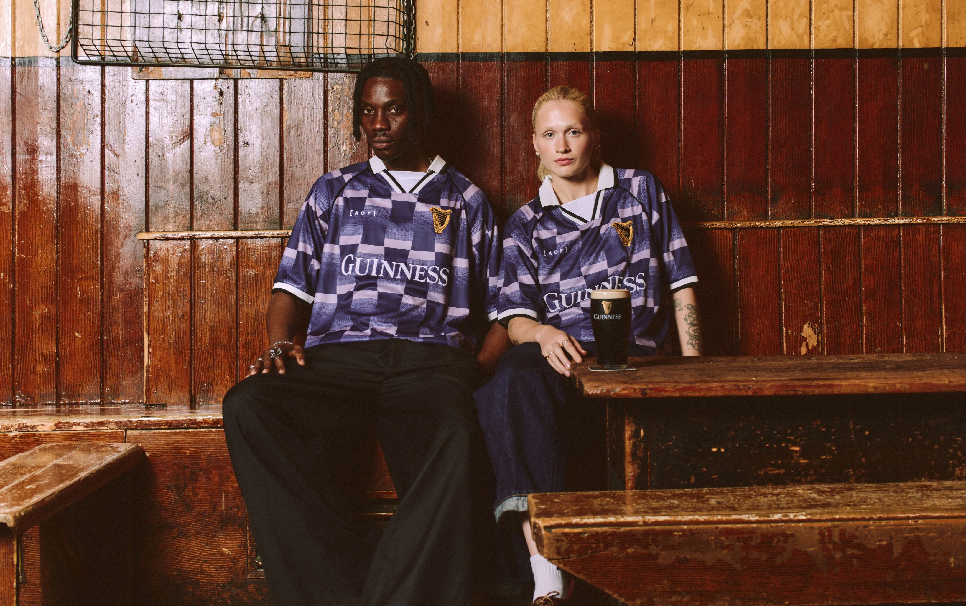 Two people in matching purple checkered Guinness shirts sit on a wooden bench in an old pub, a drink on the table in front of them.