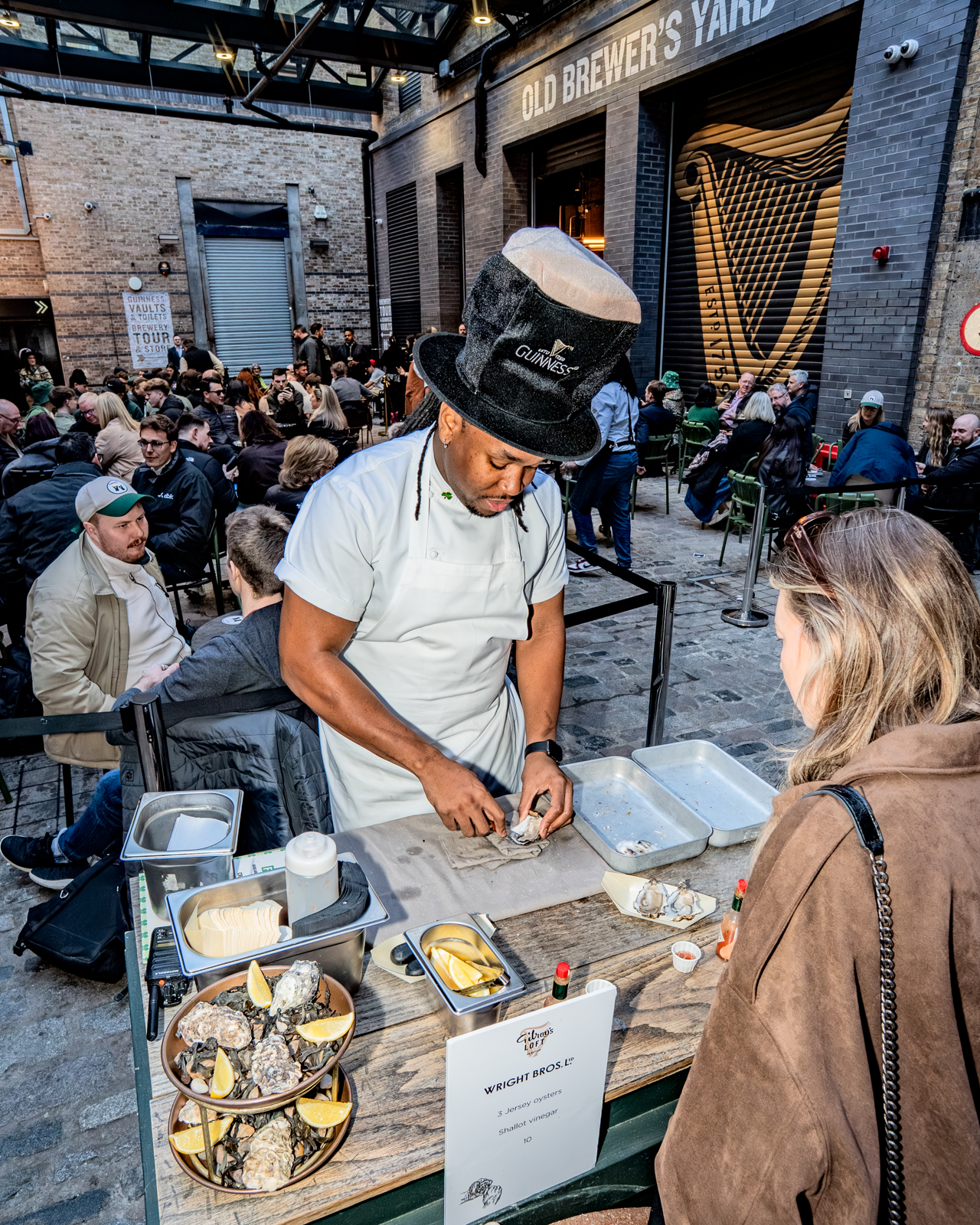 The Head Chef of Gilroy's Loft shucks an oyster in the busy beer garden courtyard in Covent Garden while people enjoy their Guinness behind him