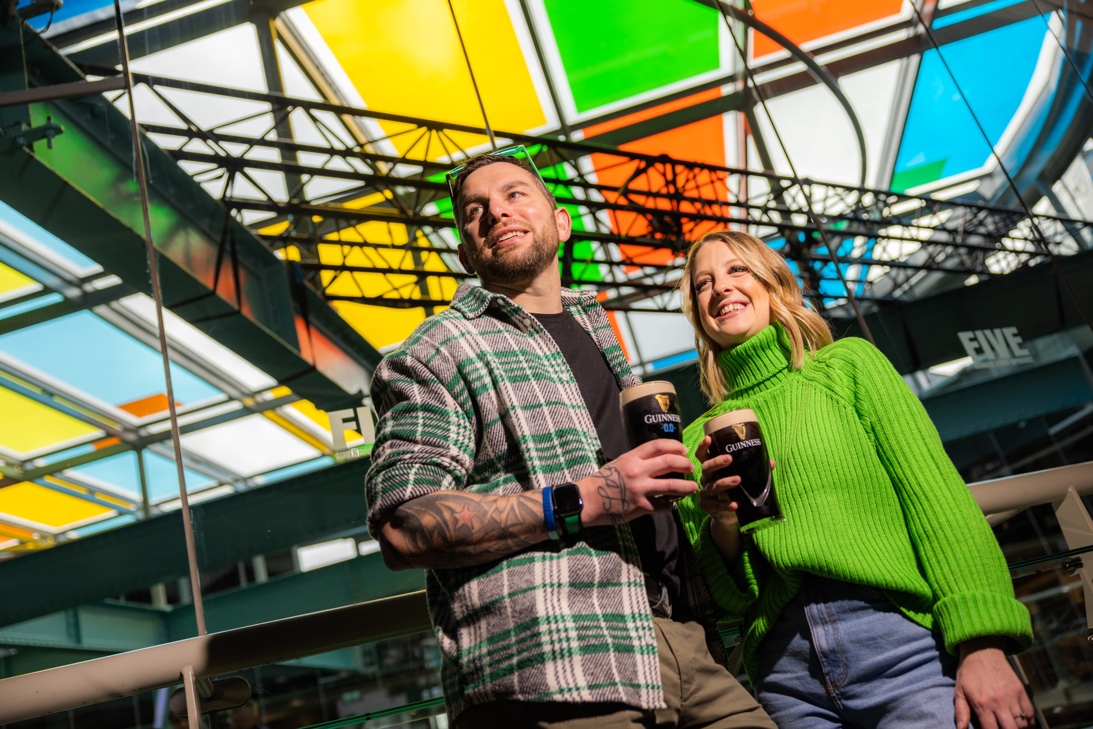 A man and a woman smile and stand outside underneath a colourful awning, both holding a pint of Guinness and wear green-hued clothing.