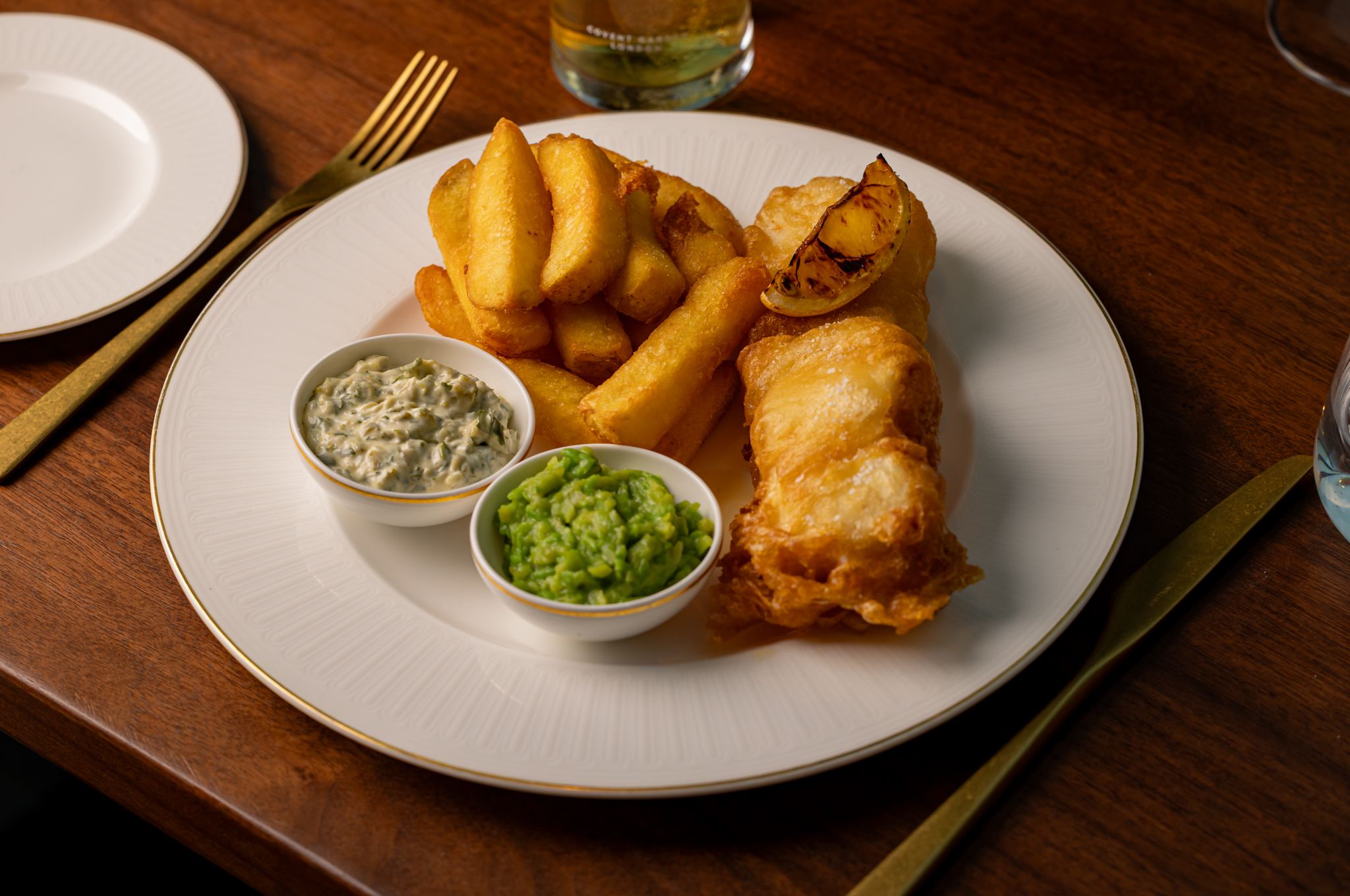 A white plate with battered fish, chunky chips and pots of mushy peas and tartar sauce