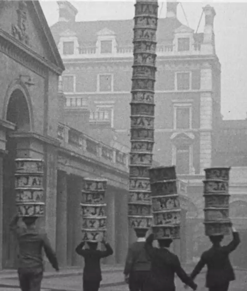 A black and white historical photo shows several people carrying tall, stacked crates on their heads in a street lined with old buildings. 