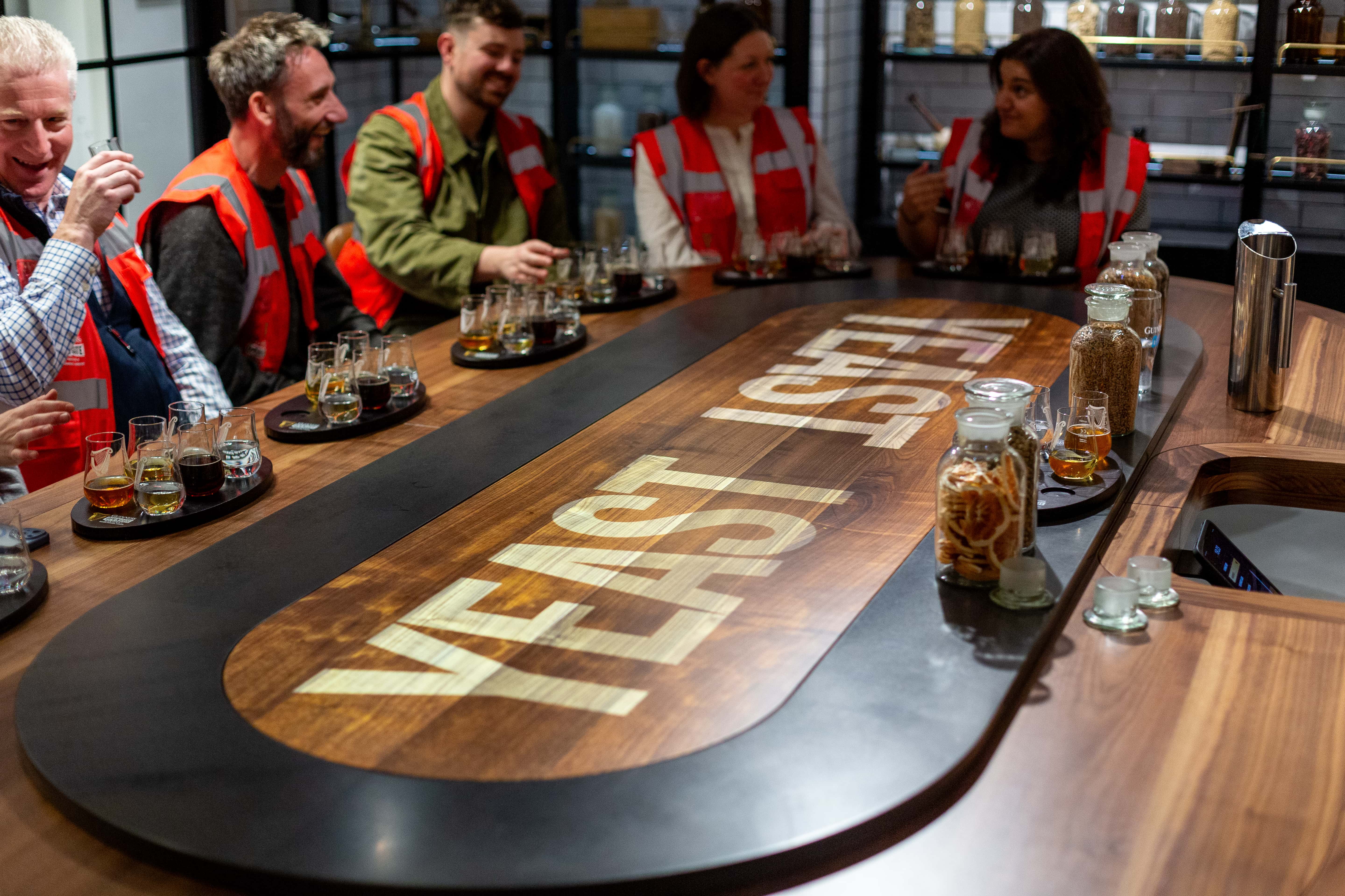 A tour group sit smiling around a table in the Brewer's Room on the Open Gate Experience in Covent Garden, tasting the craft beers made in the brewery