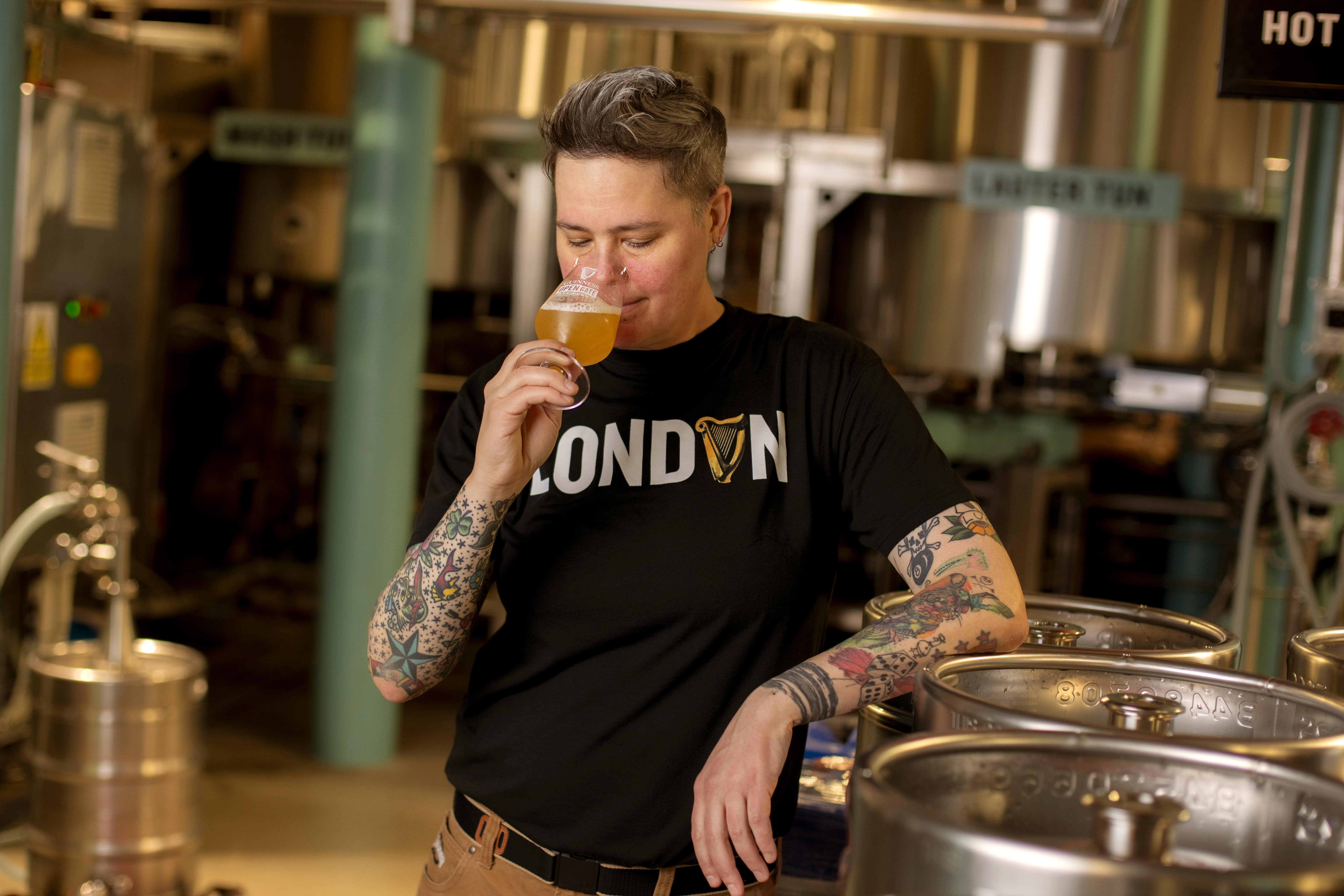 Master Brewer Hollie Stephenson leans against a metal beer keg in the brewery and holds a glass of beer to her nose.