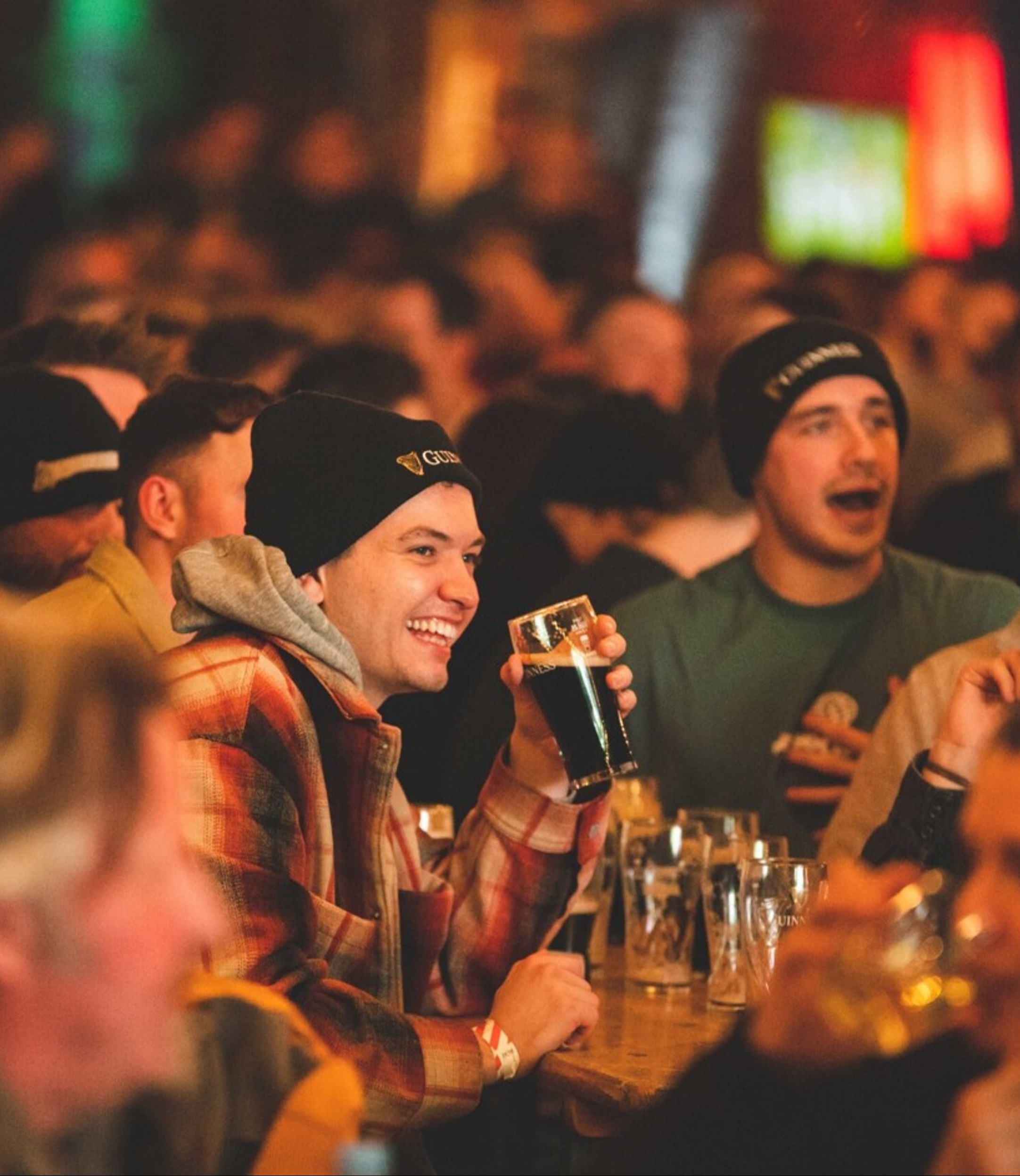 Smiling men in beanies drink dark beer at a lively bar, with other people enjoying themselves in the bustling background. 