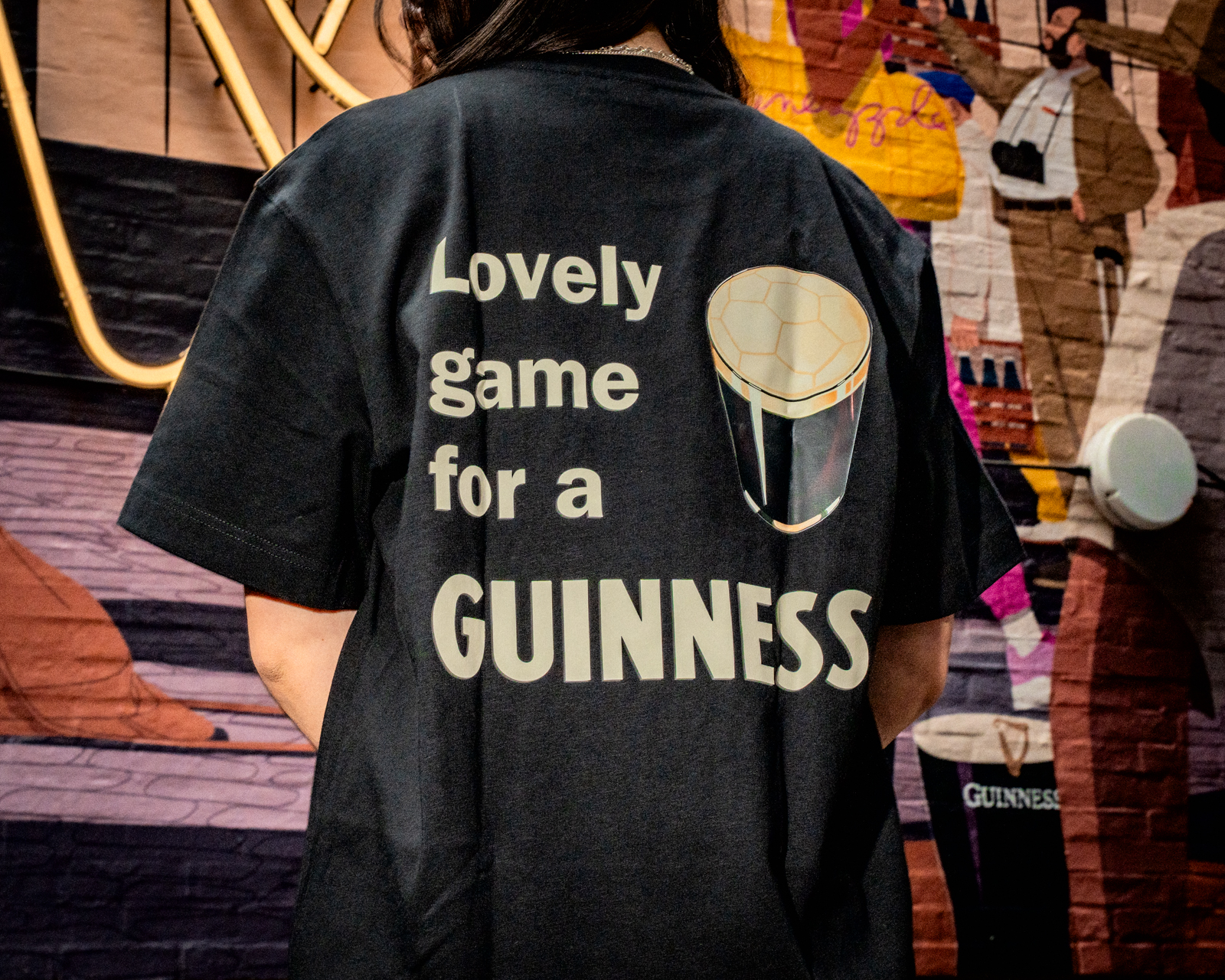 A person with their back to the camera wears a black T shirt with Lovely Game for a Guinness inscribed on the back with a Guinness pint graphic