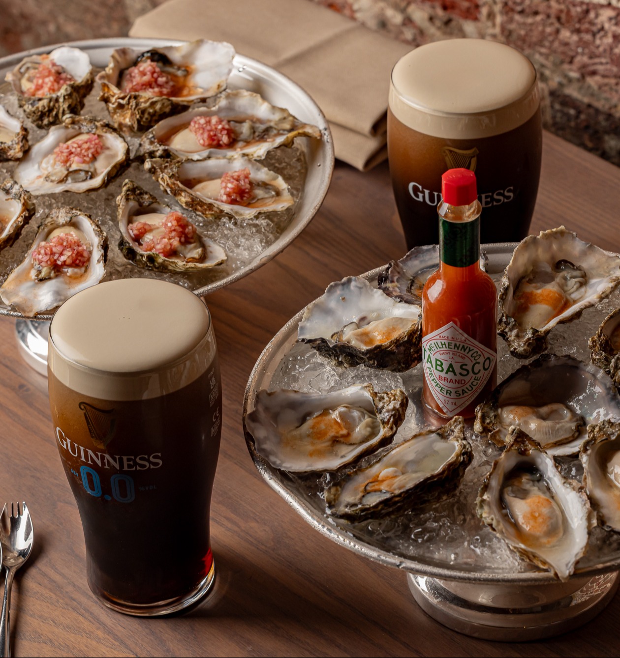 Two plates of oysters on ice, some topped with red sauce, are on a wooden table alongside two glasses of Guinness and a bottle of Tabasco. 