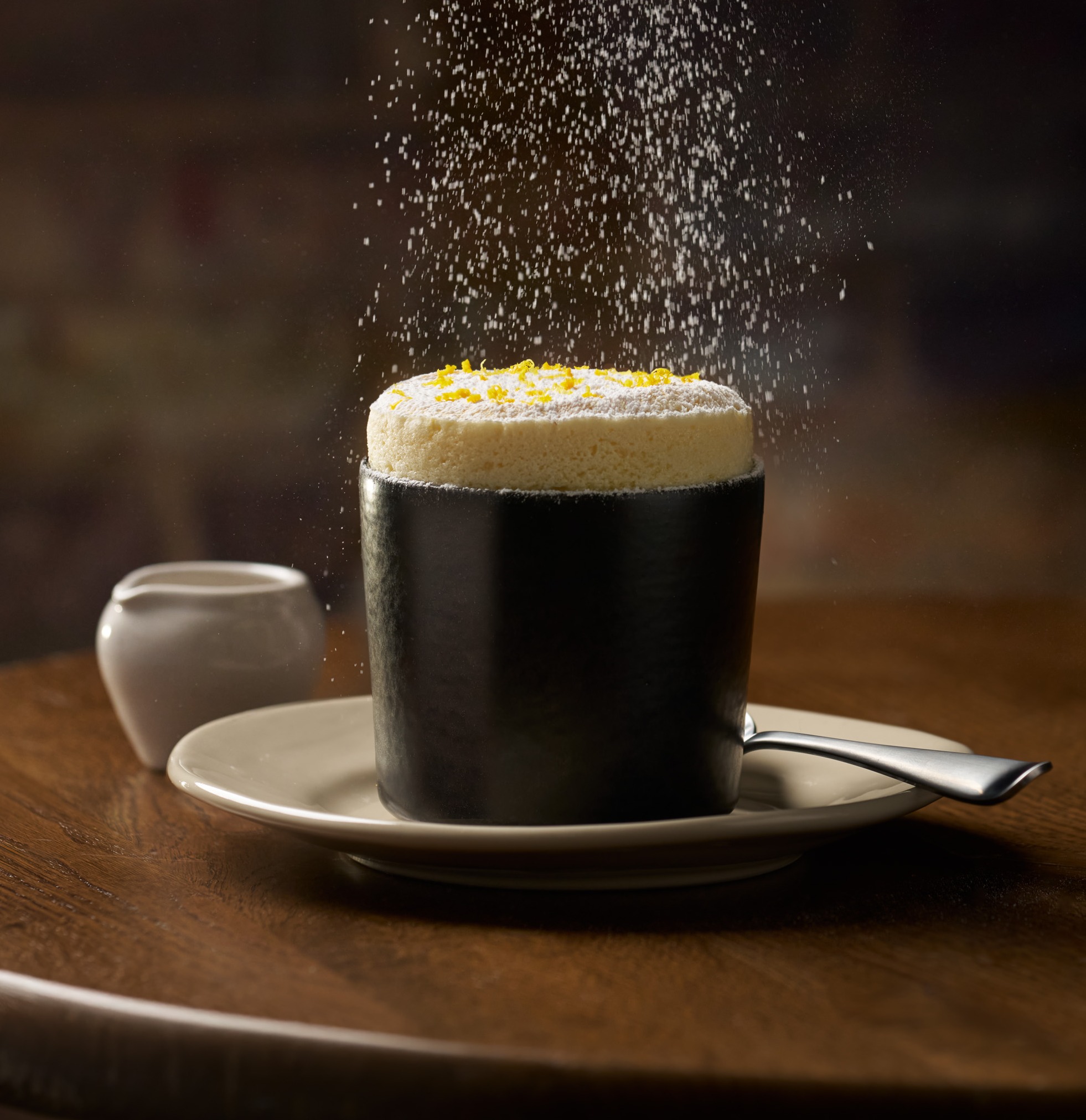 A dark mug with a frothy, white-topped drink, garnished with lemon zest, sprinkled with white powder, on a wooden table. 