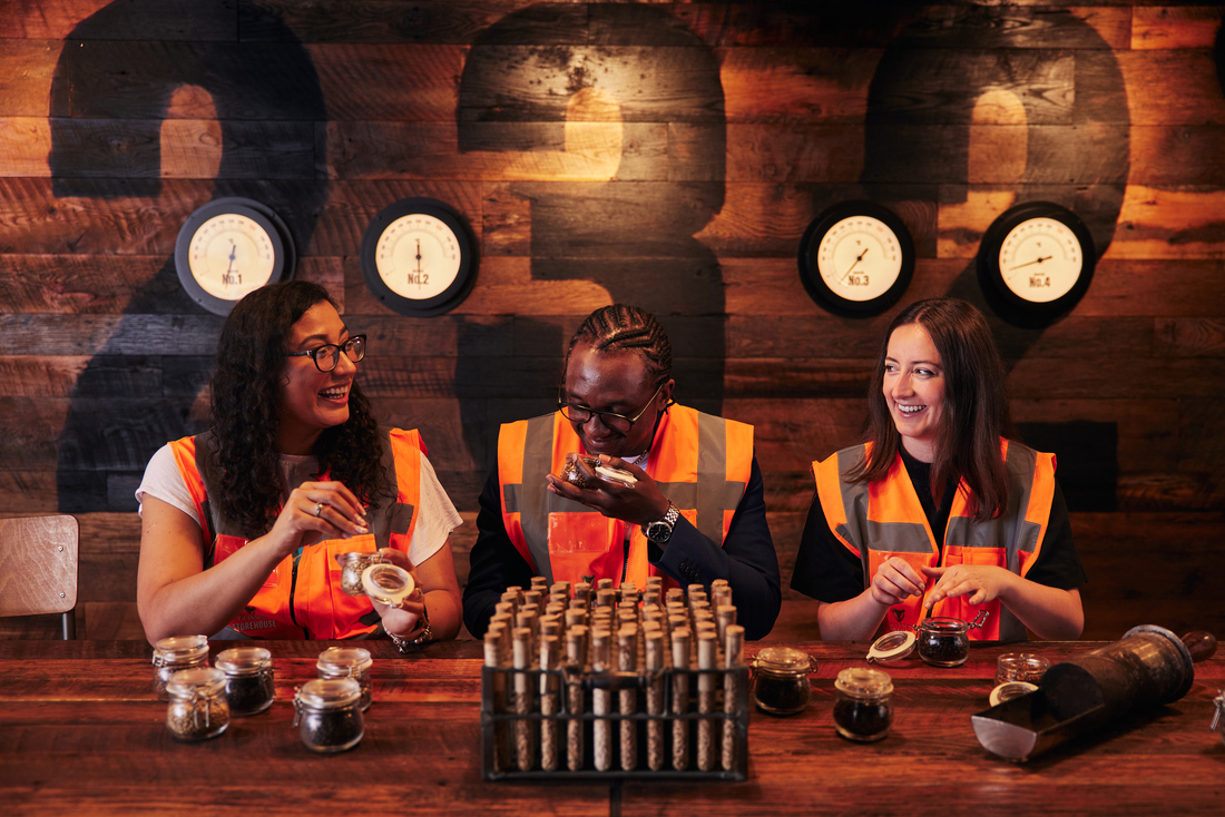 Three women sit behind an array of test tubes. They are smelling small glass jars and laughing.