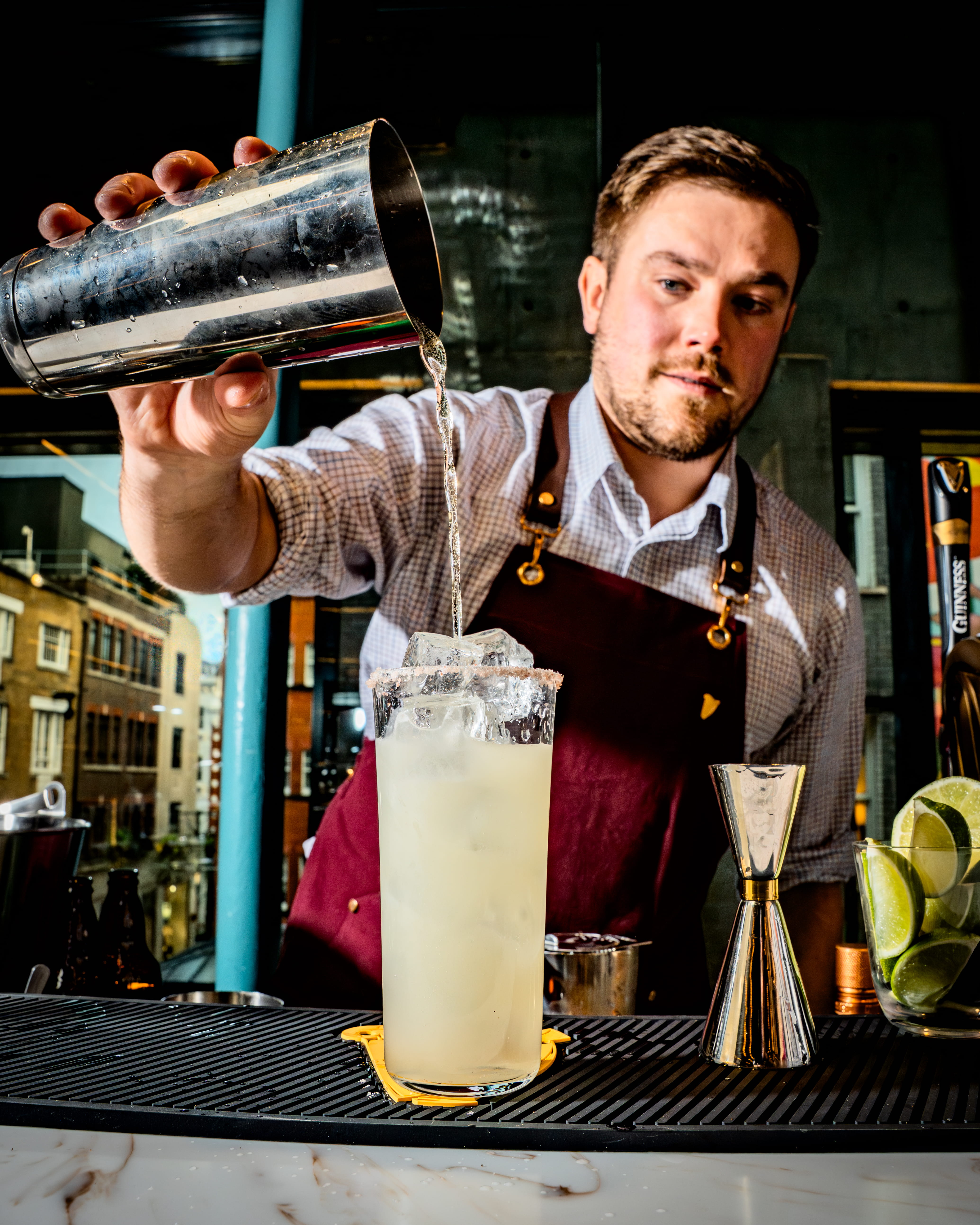 A bartender carefully pours a freshly shaken margarita into a tall, iced glass with a salt rim. Adding crisp lager to create a lagerita cocktail.