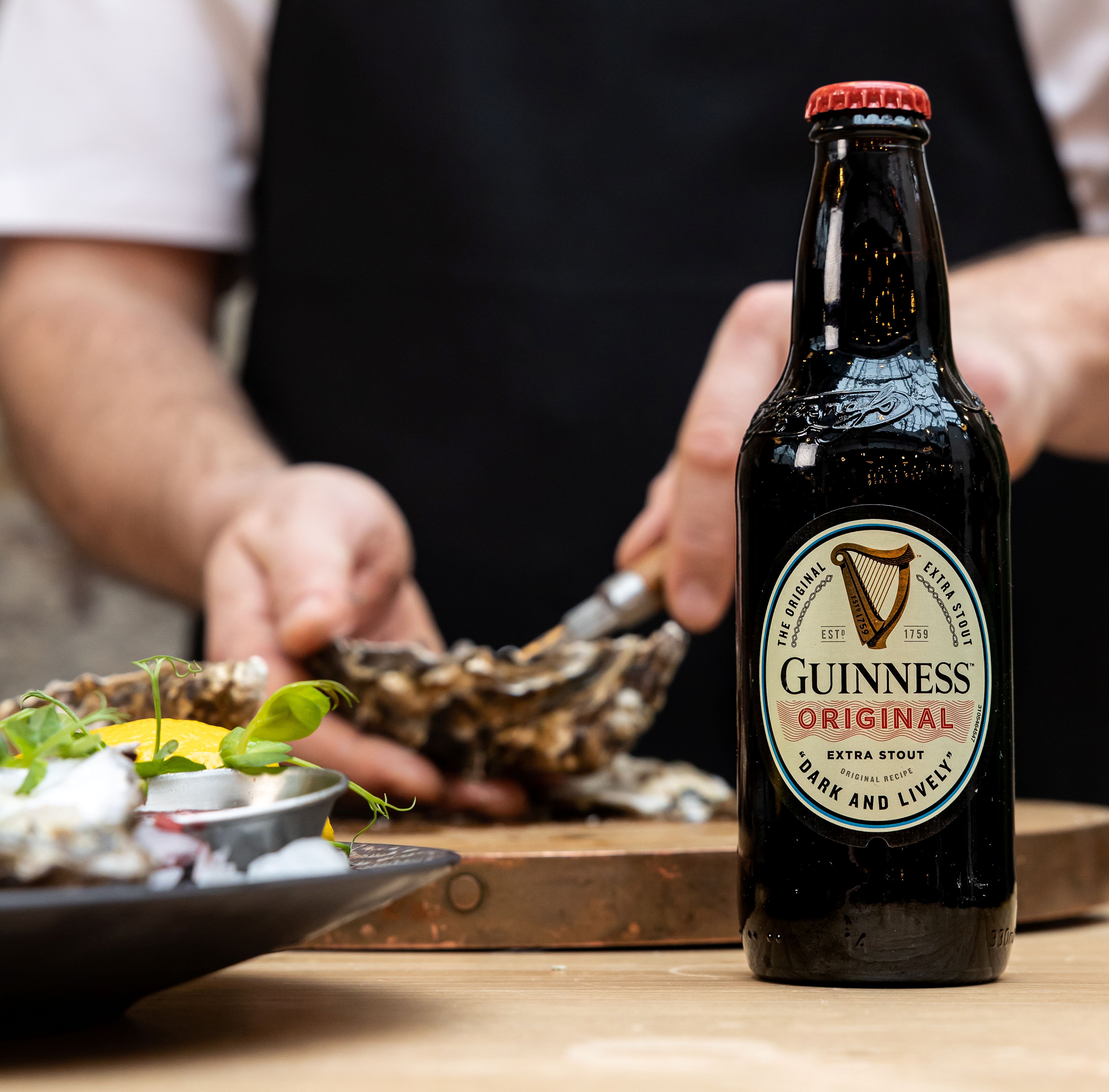 A dish on a plate, Guinness Original next to it. Chef is preparing next meal in a background.
