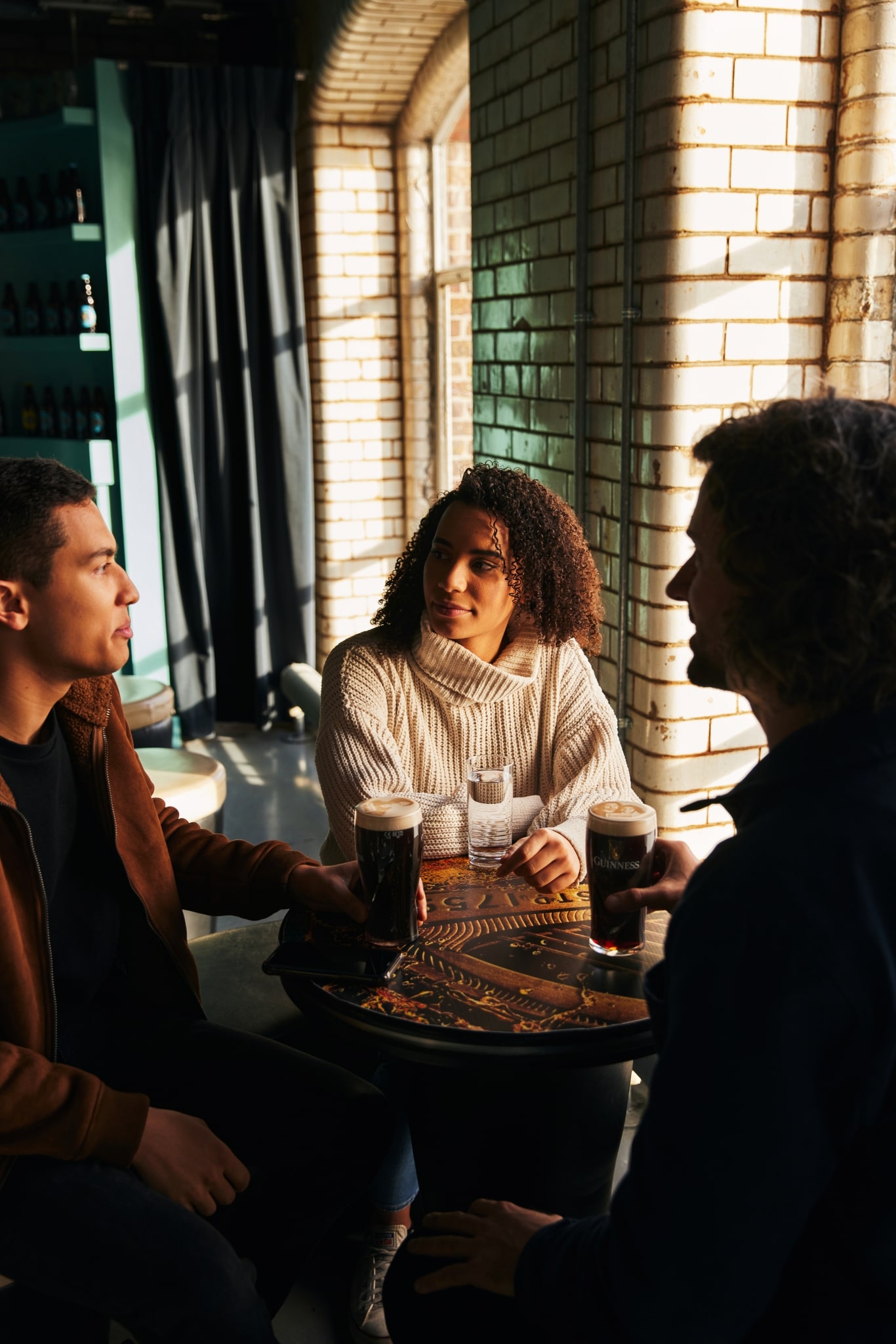 Three people sit around a table, tow have pints of Guinness and the sun streams in through the blinds.