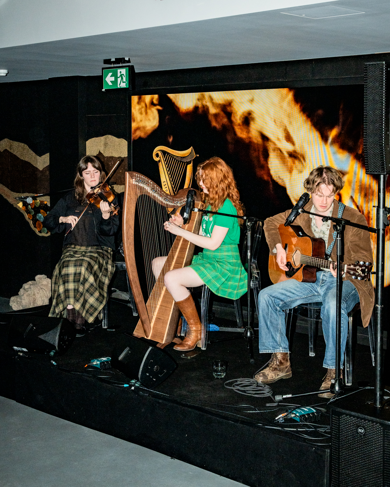 Three musicians sat down on stage playing instruments, a harp, violin and guitar with a Guinness logo in the background