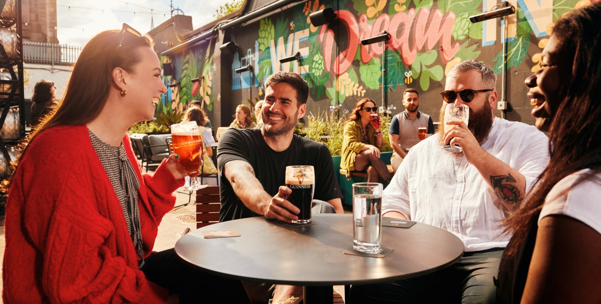 Smiling friends enjoying Guinness and other drinks at an outdoor table with a vibrant mural in the background