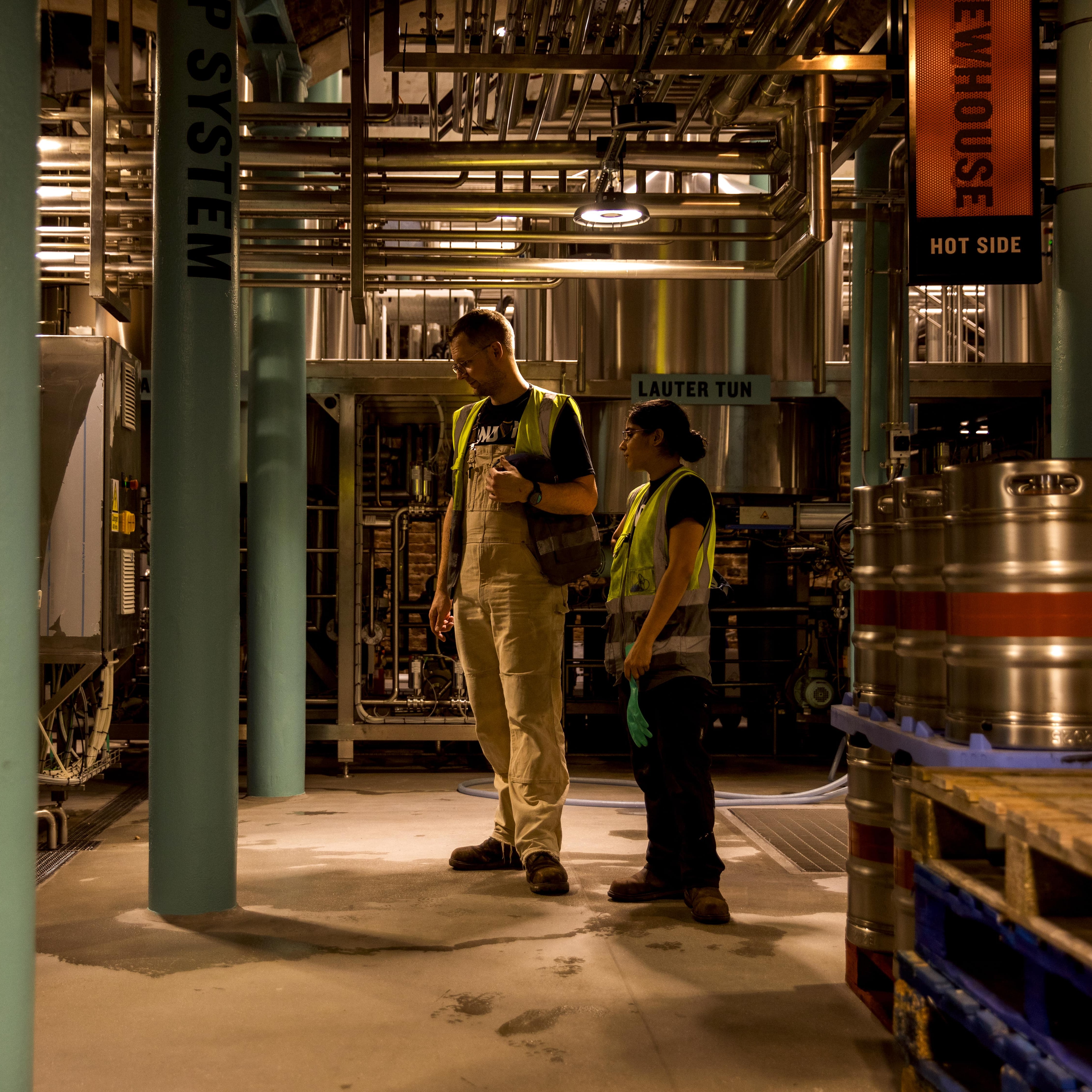 Two brewers, a man and a woman, with safety jackets on stand and watch the brewing equipment in the micro-brewery