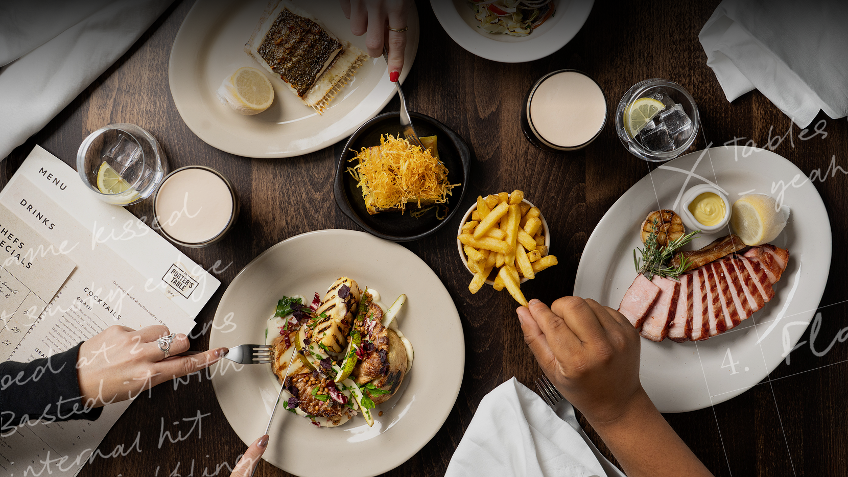 Overhead shot of a wooden dining table with plates of grilled fish, sliced meat, and fries. Drinks, a menu, and hands holding cutlery for dining 