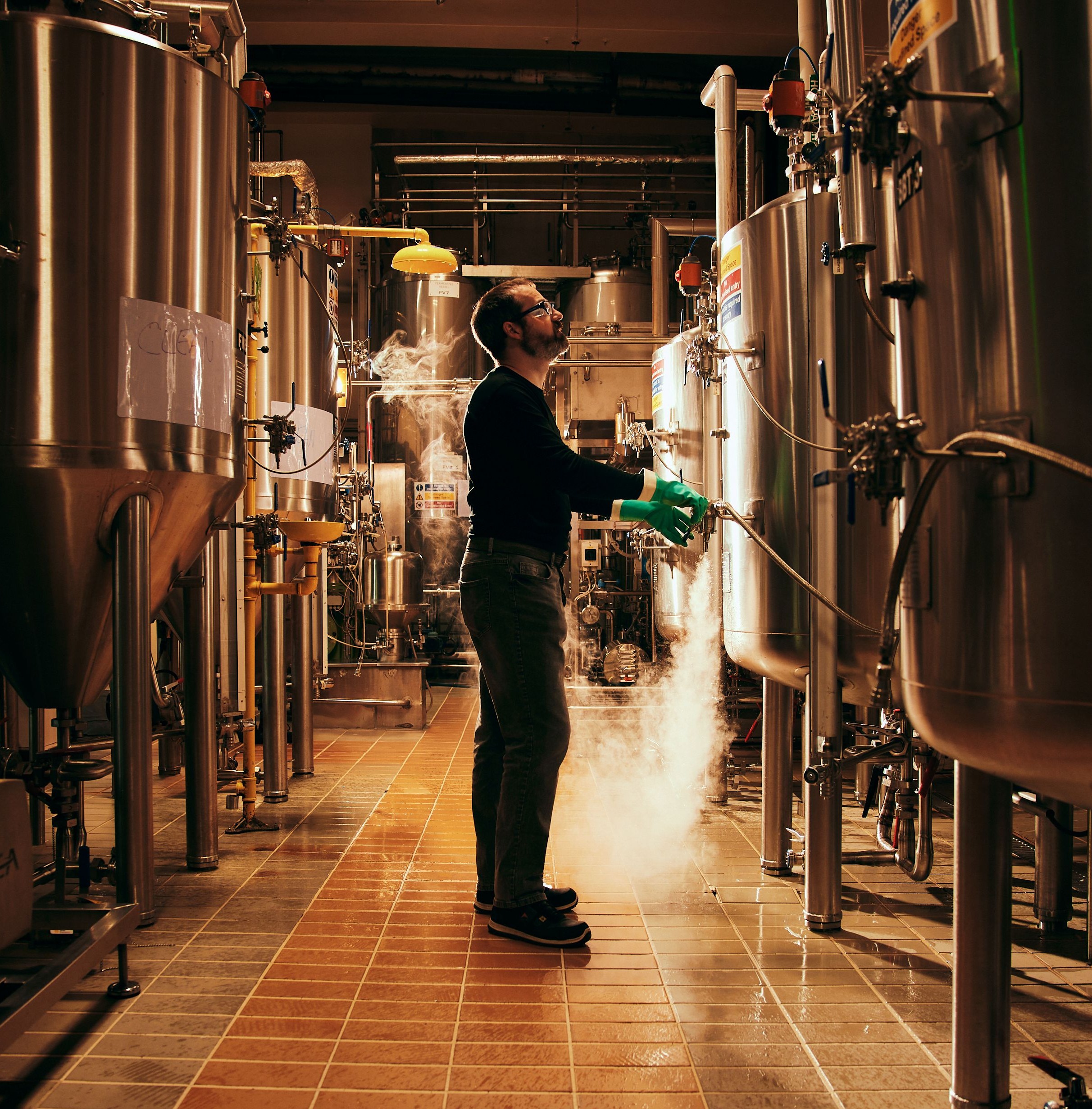 A man in green gloves stands amidst large stainless steel tanks in a brewery, with steam rising from one tank.