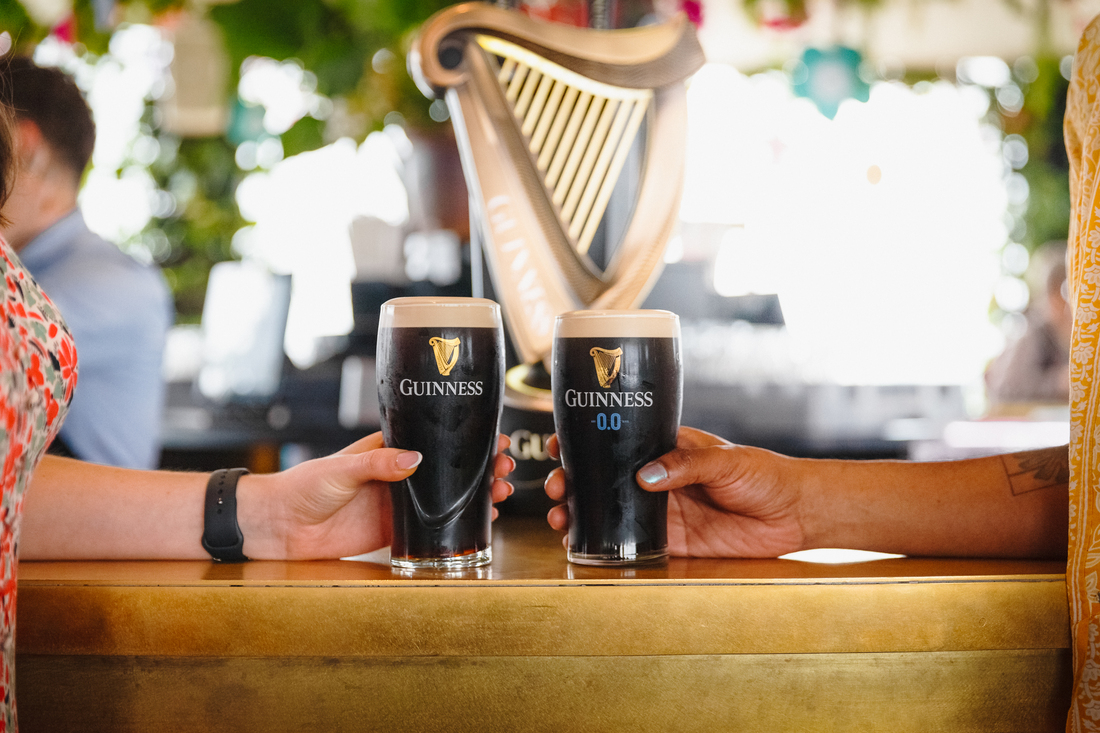 2 hands holding 2 pints of Guinness. One is holding Guinness poured in Guinness glass and the second is holding a Guinness in Guinness 0.0 glass