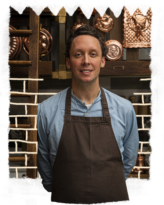 A smiling man in a brown apron, with copper decorations on shelves and a brick wall in the background.