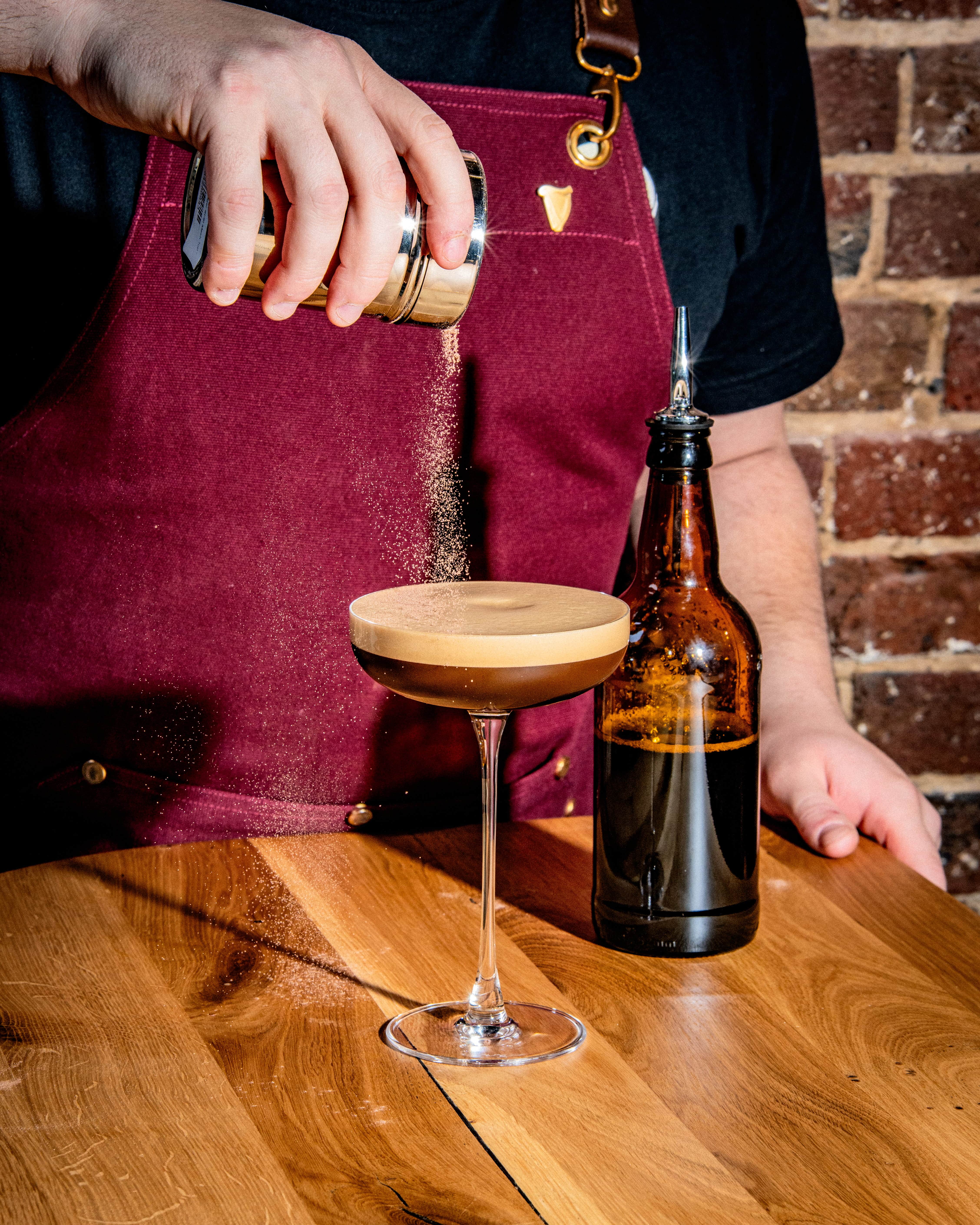 A cocktail on a wooden table in a martini glass, with a bartender sprinkling chocolate on top