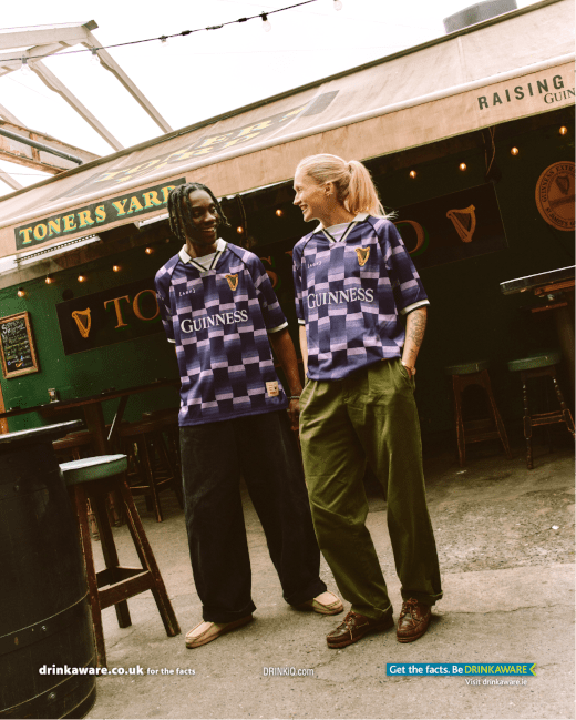 Two people in purple patterned shirts stand in a pub with a Guinness sign, smiling at each other. 