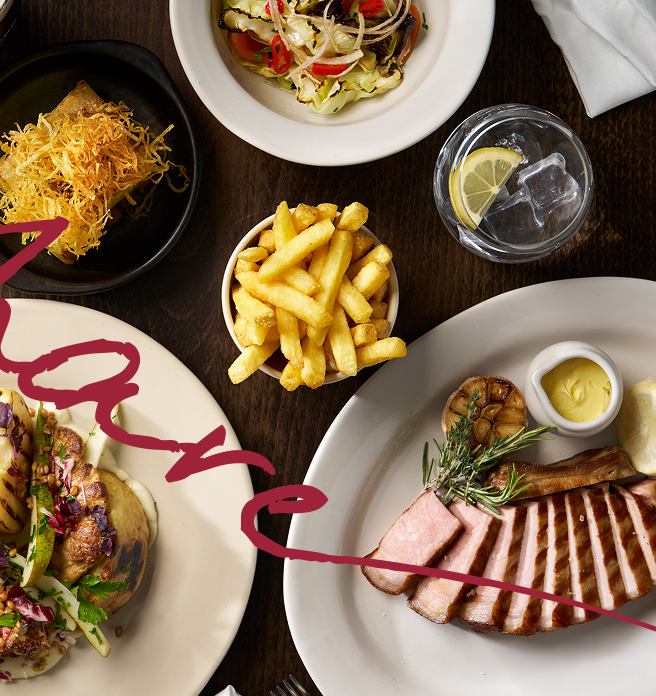 Dark wooden table with sliced meat and rosemary, french fries, salad, shredded crispy food, and a glass of water with lemon. 