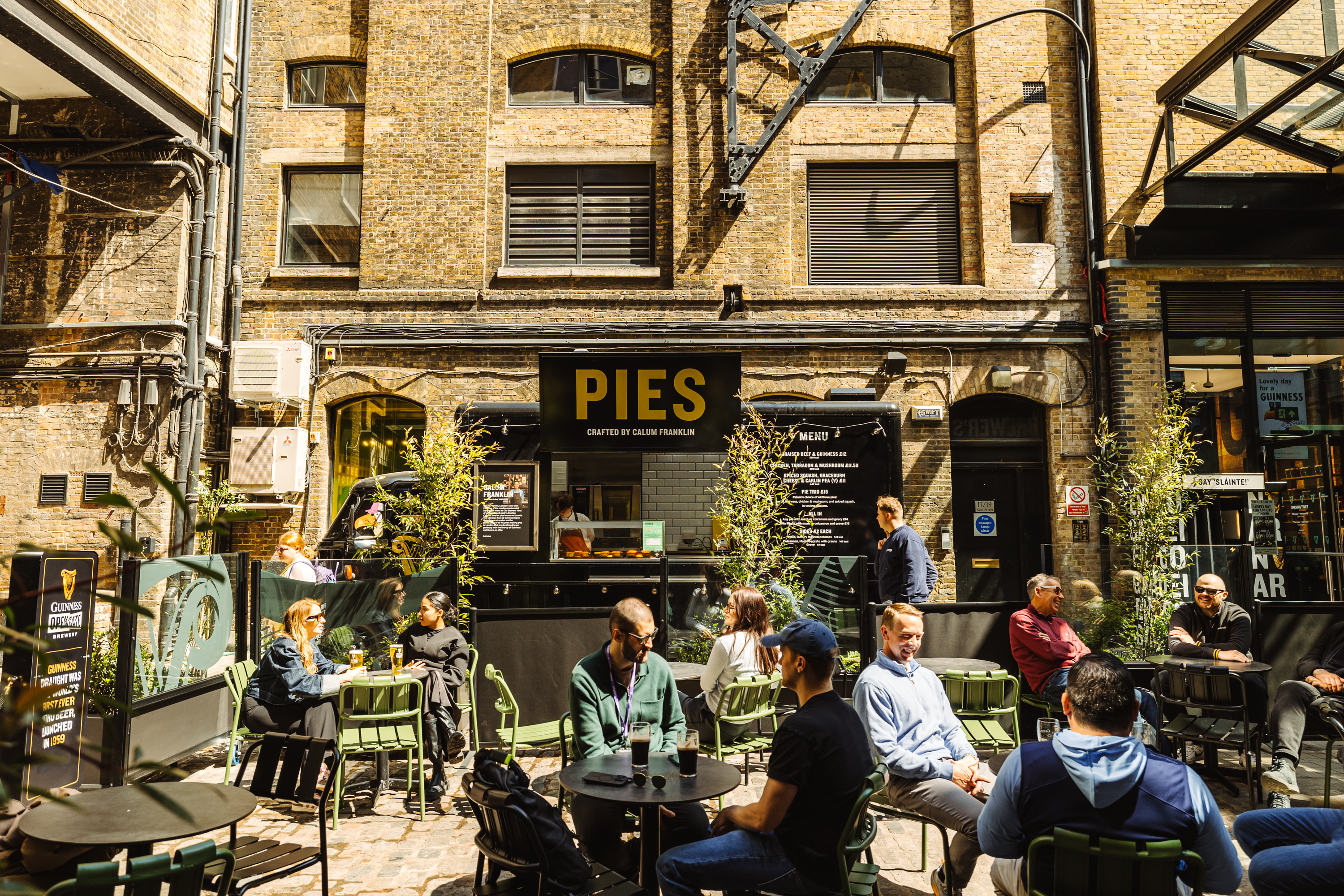 A pub courtyard with multiple tables and people drinking with a van selling pies in the background