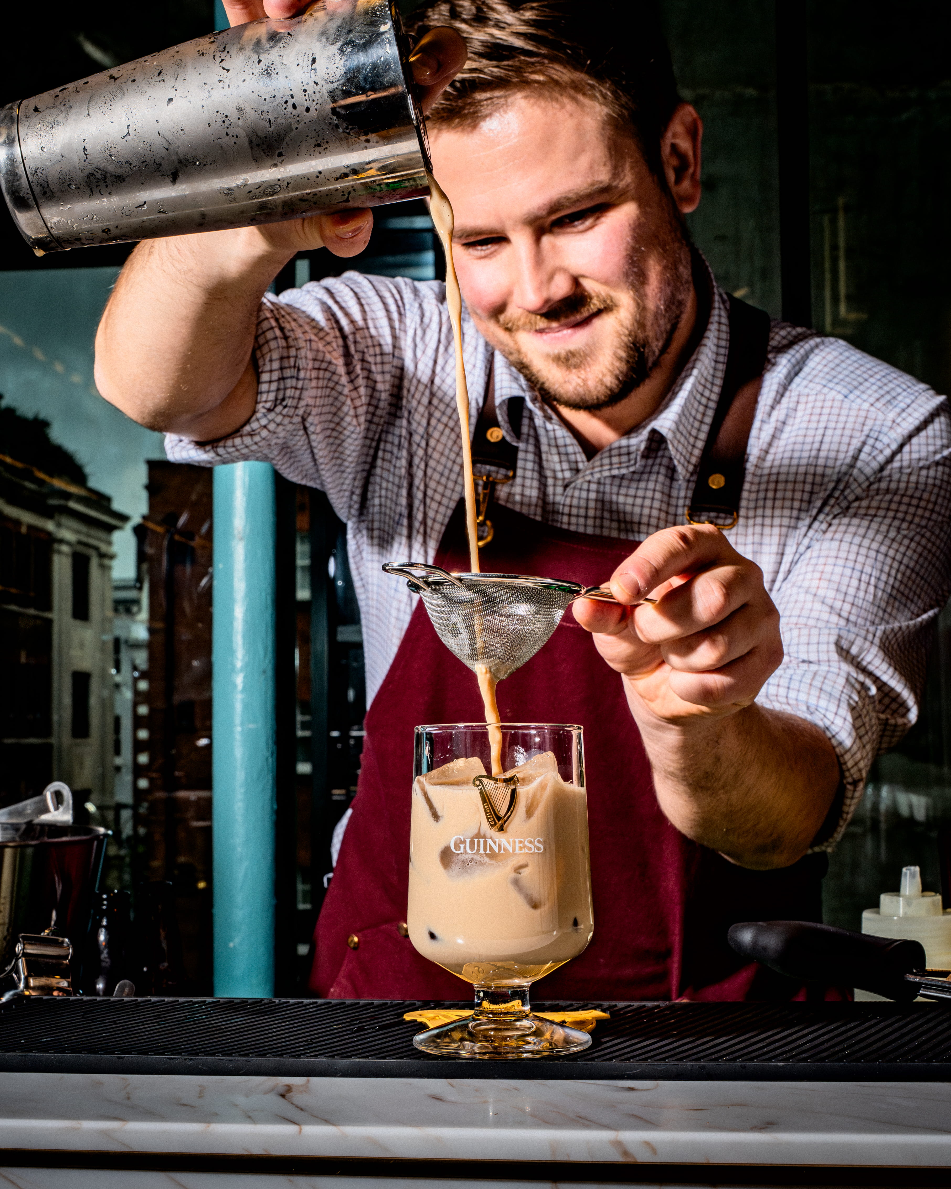 A bartender pours a freshly shaken cocktail from the shaker and strains it into a tall glass filled with ice.