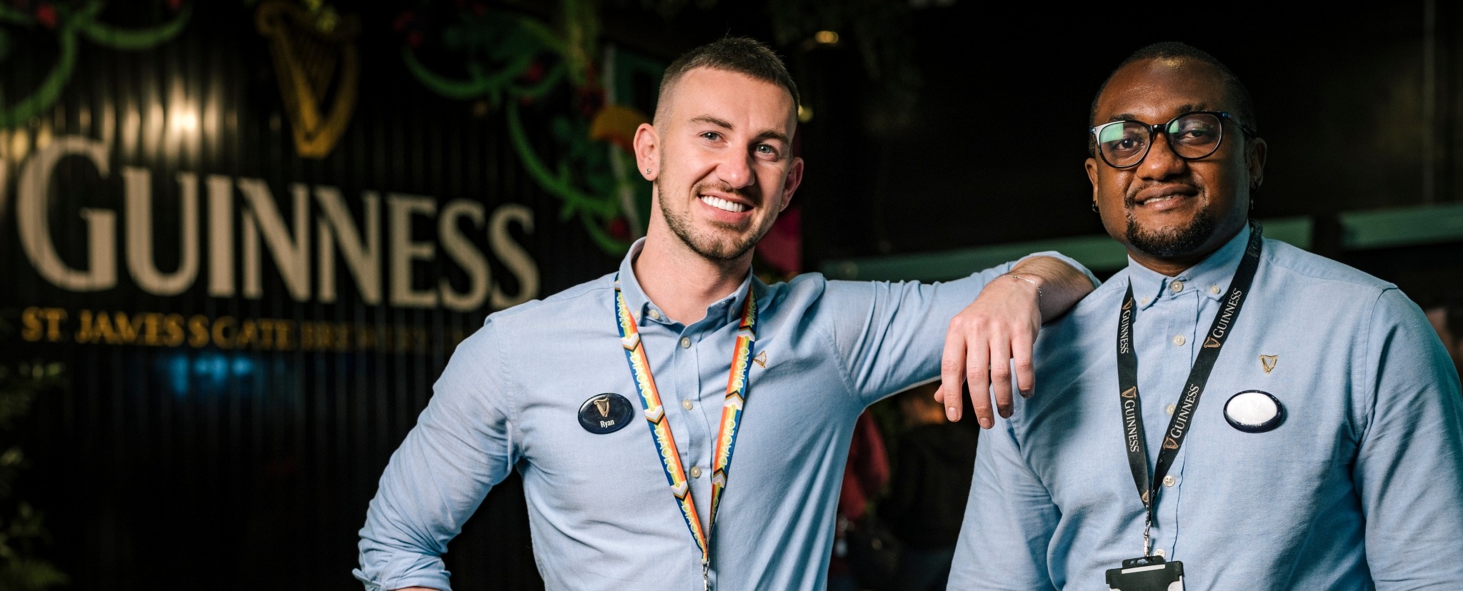 Two men in blue shirts face the camera smiling. They both wear Guinness name badges and lanyards.