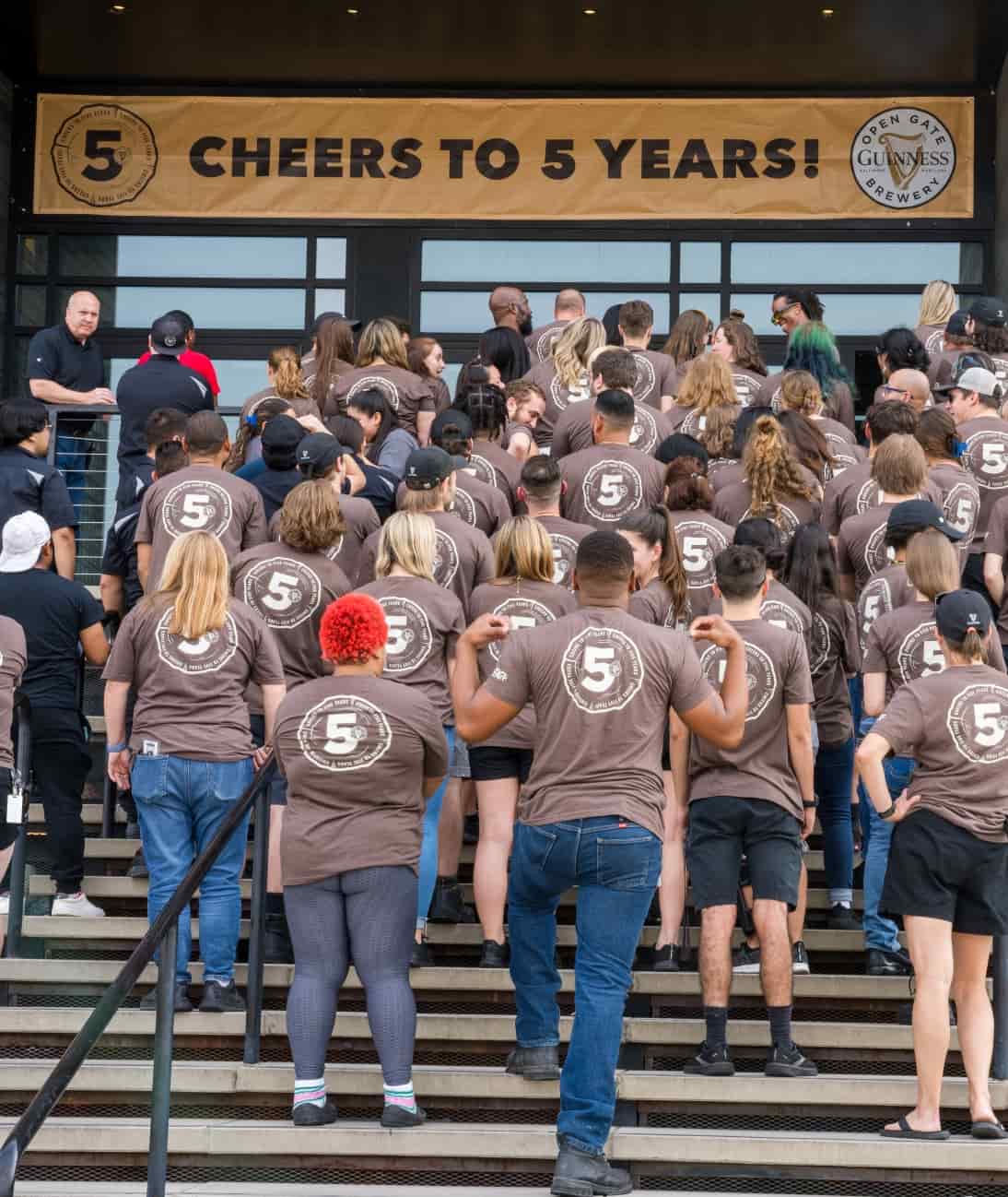 A large group of people wearing the same tshirt stand on some steps under a banner that reads "cheers to 5 years."