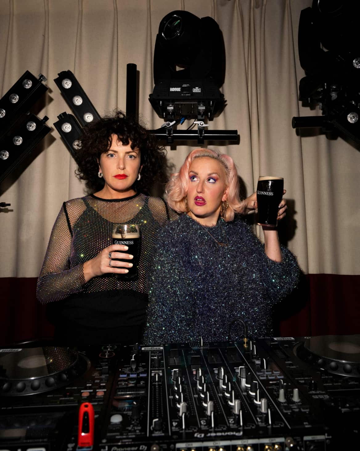 Annie Mac and Aimee Phillips stand behind a DJ deck holding pints of Guinness.