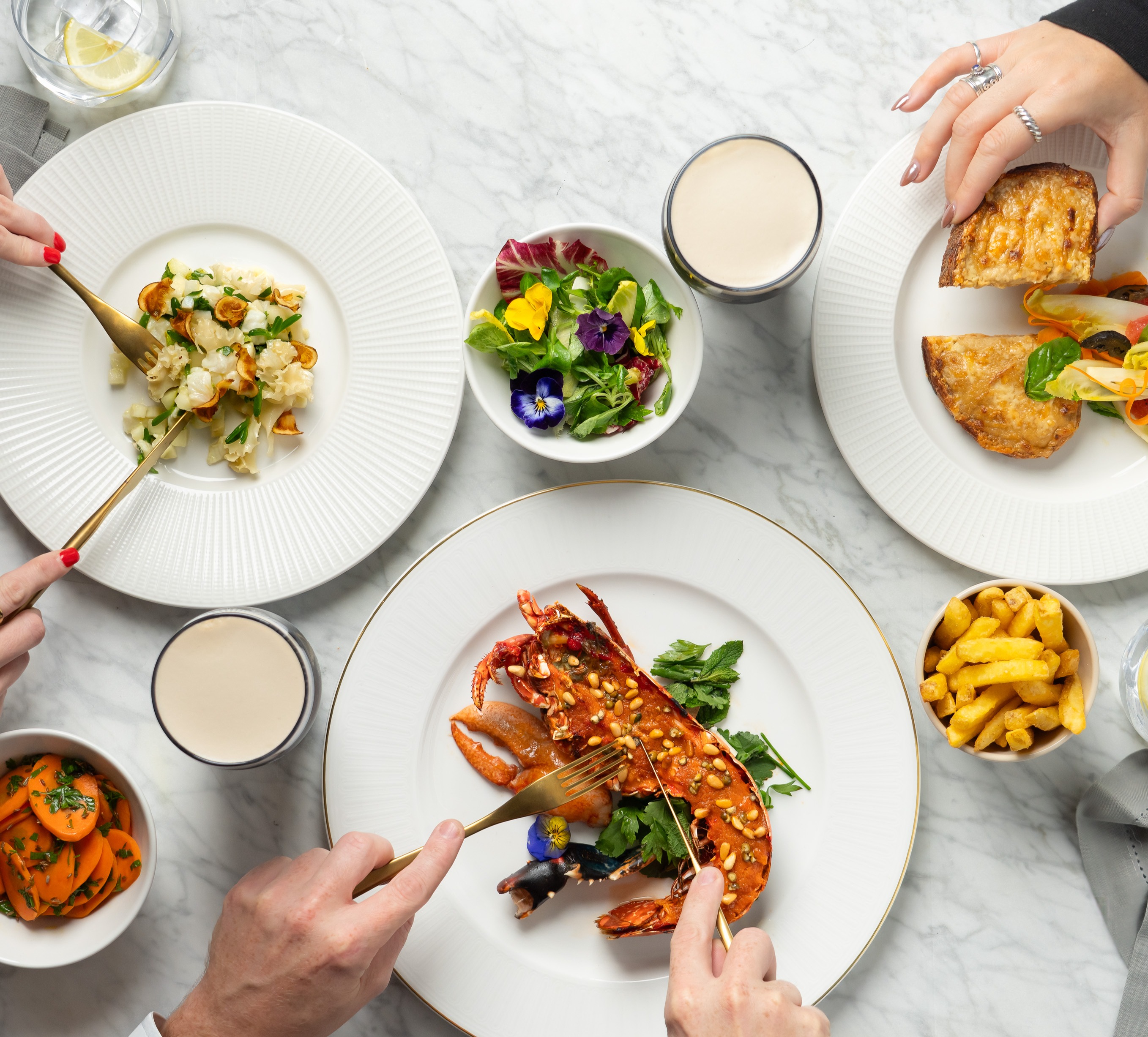 A top-down view of several people dining at a marble table, enjoying dishes like a whole lobster, cauliflower, pastries, salad, carrots, and fries.