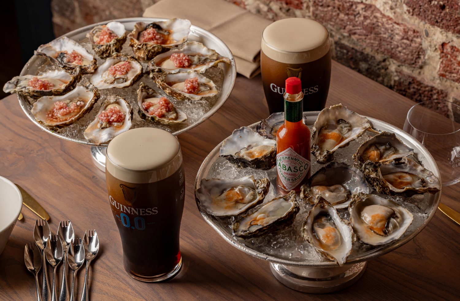 Two plates of oysters on ice, some topped with red sauce, are on a wooden table alongside two glasses of Guinness and a bottle of Tabasco. 