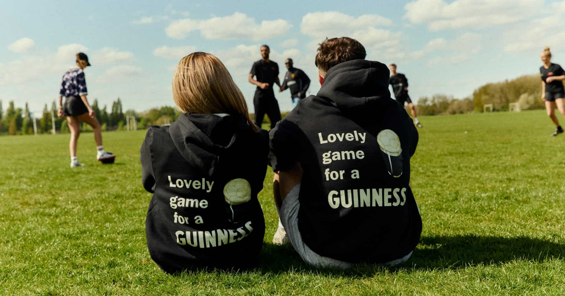 Two people sit on grass facing away from the camera, both wearing Guinness hoodies. They watch people playing football.