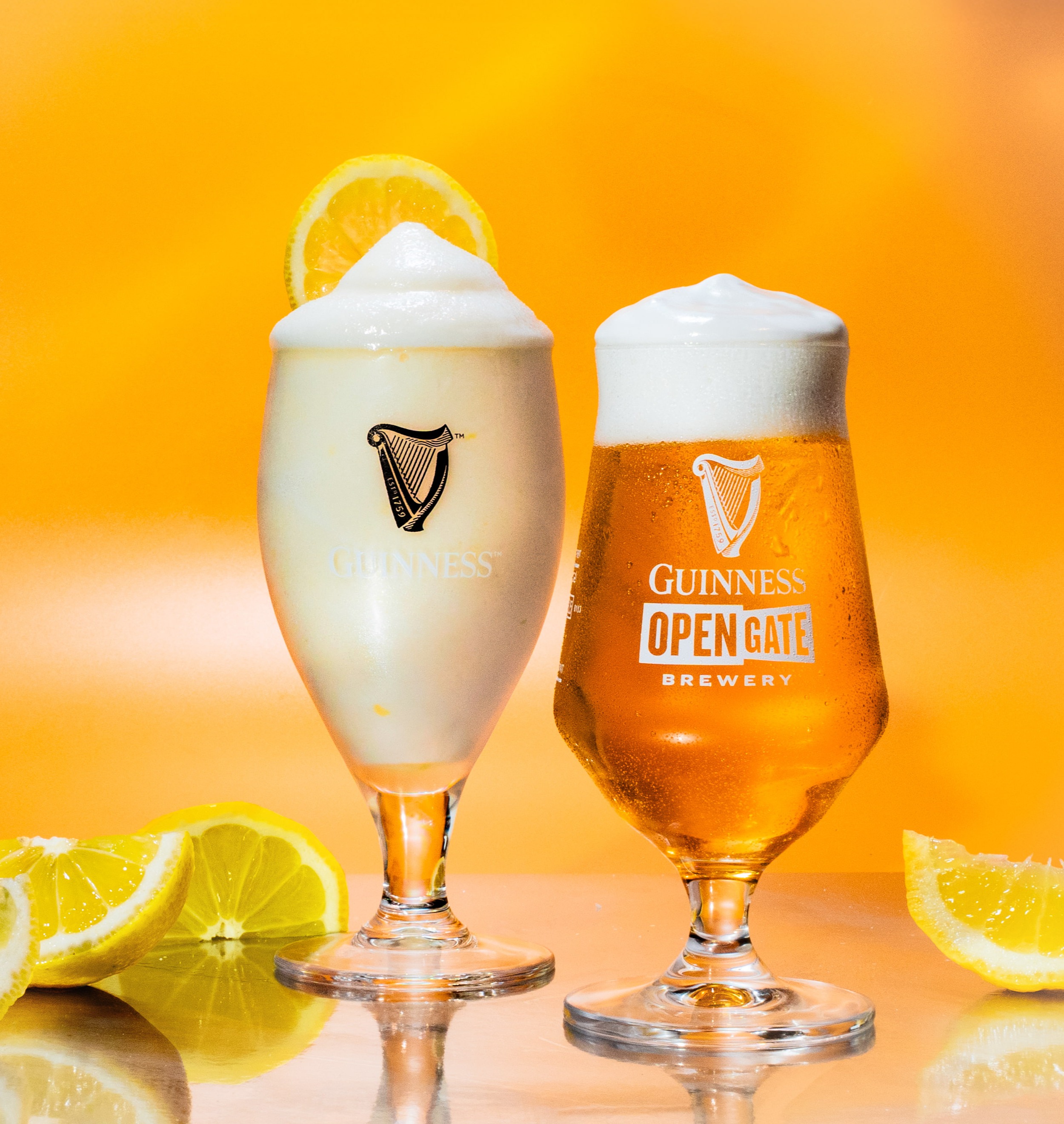 A half pint of beer with a foamy head and an iced slushy beer in Guinness glasses and slices of lemon against a yellow background