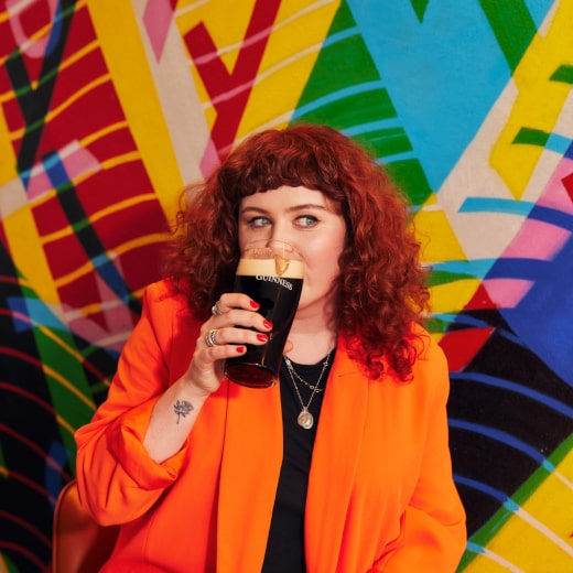 A woman with red, curly hair, wearing an orange jacket, sips from a dark pint glass against a vibrant, colorful geometric background. 