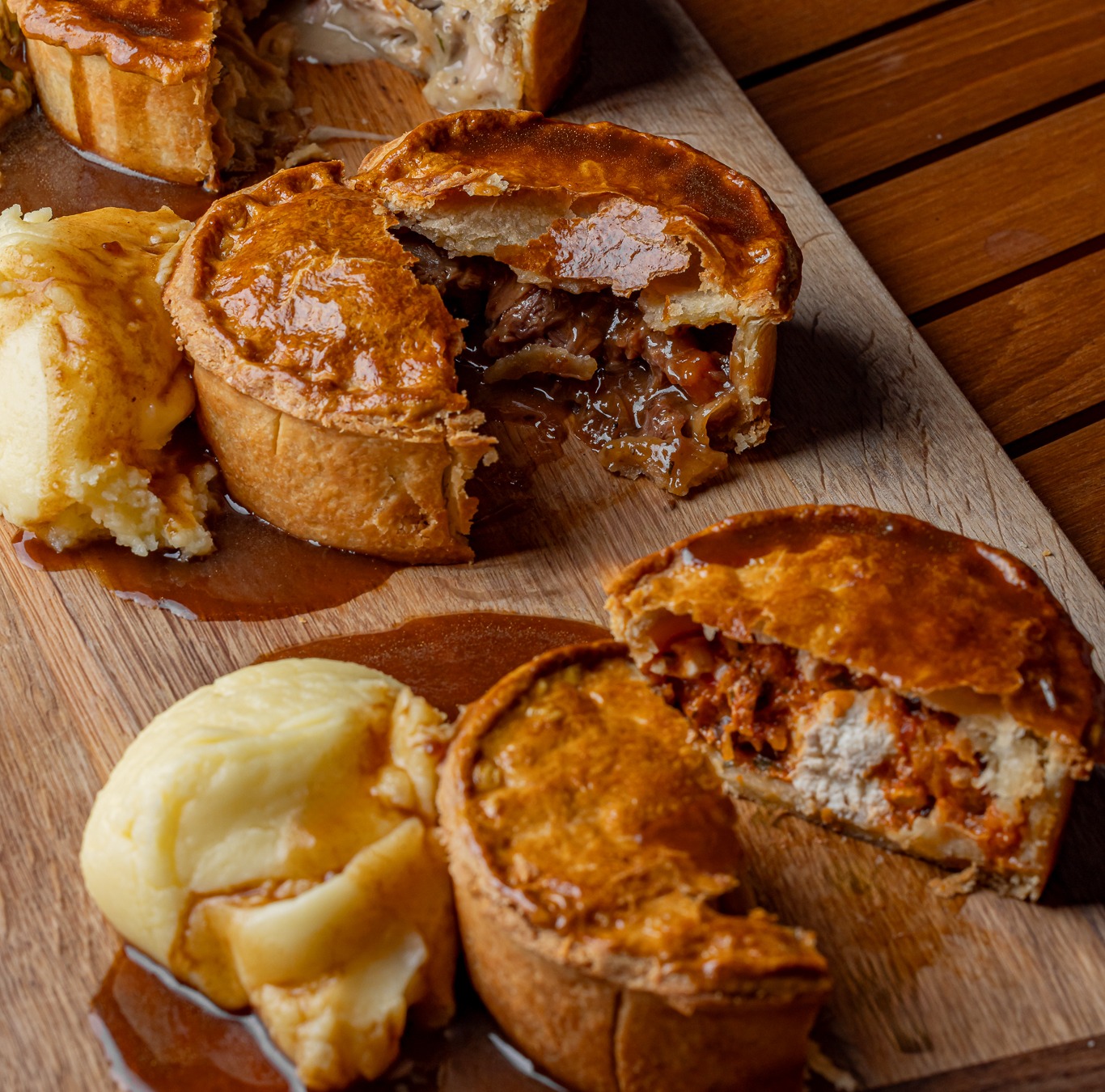 Several golden-brown savory pies, cut open to show creamy, beef, and reddish shredded meat fillings, with mashed potatoes and gravy on a wooden board.