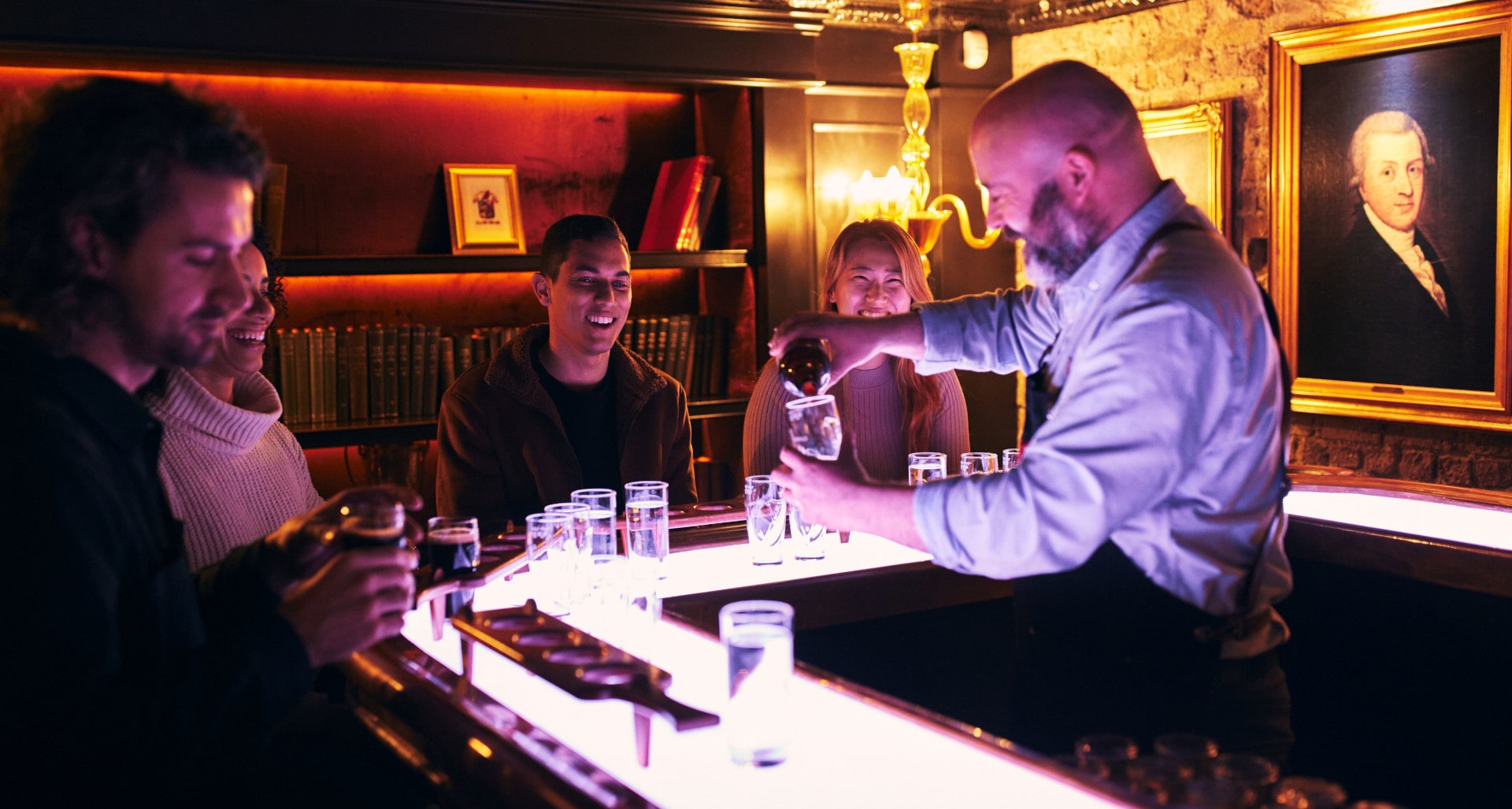 A bartender pours Guinness into a glass for smiling guests at a modern, brightly lit bar counter.  