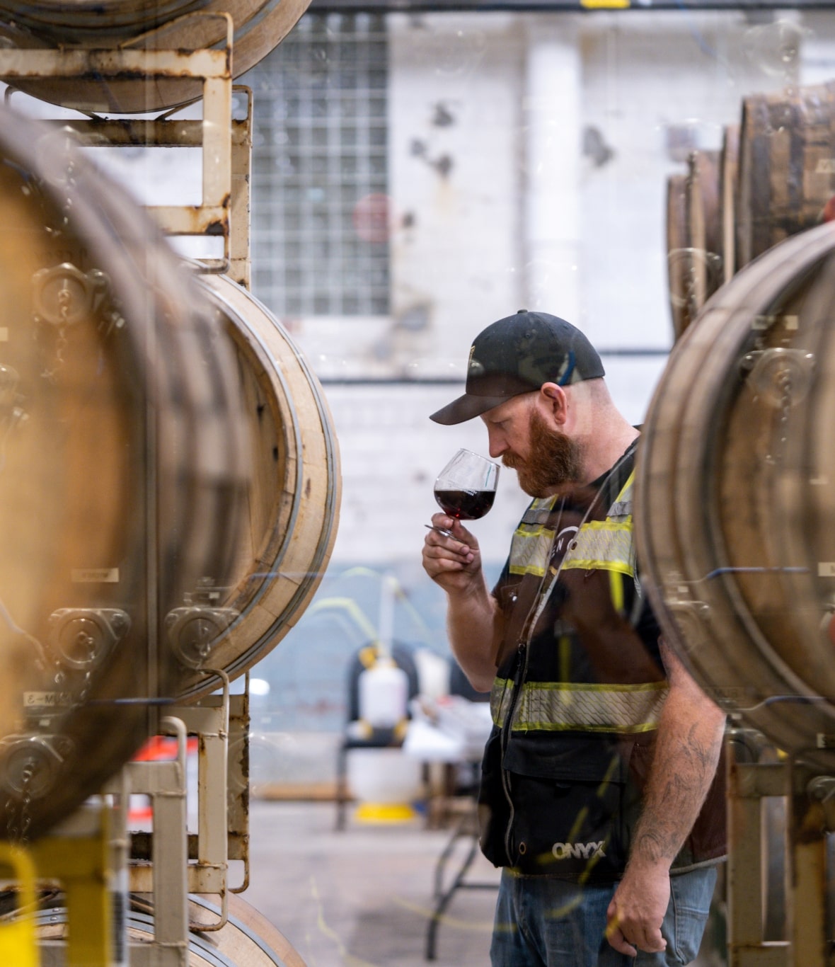 A brewery worker stands amid barrels sniffing a small glass of beer.