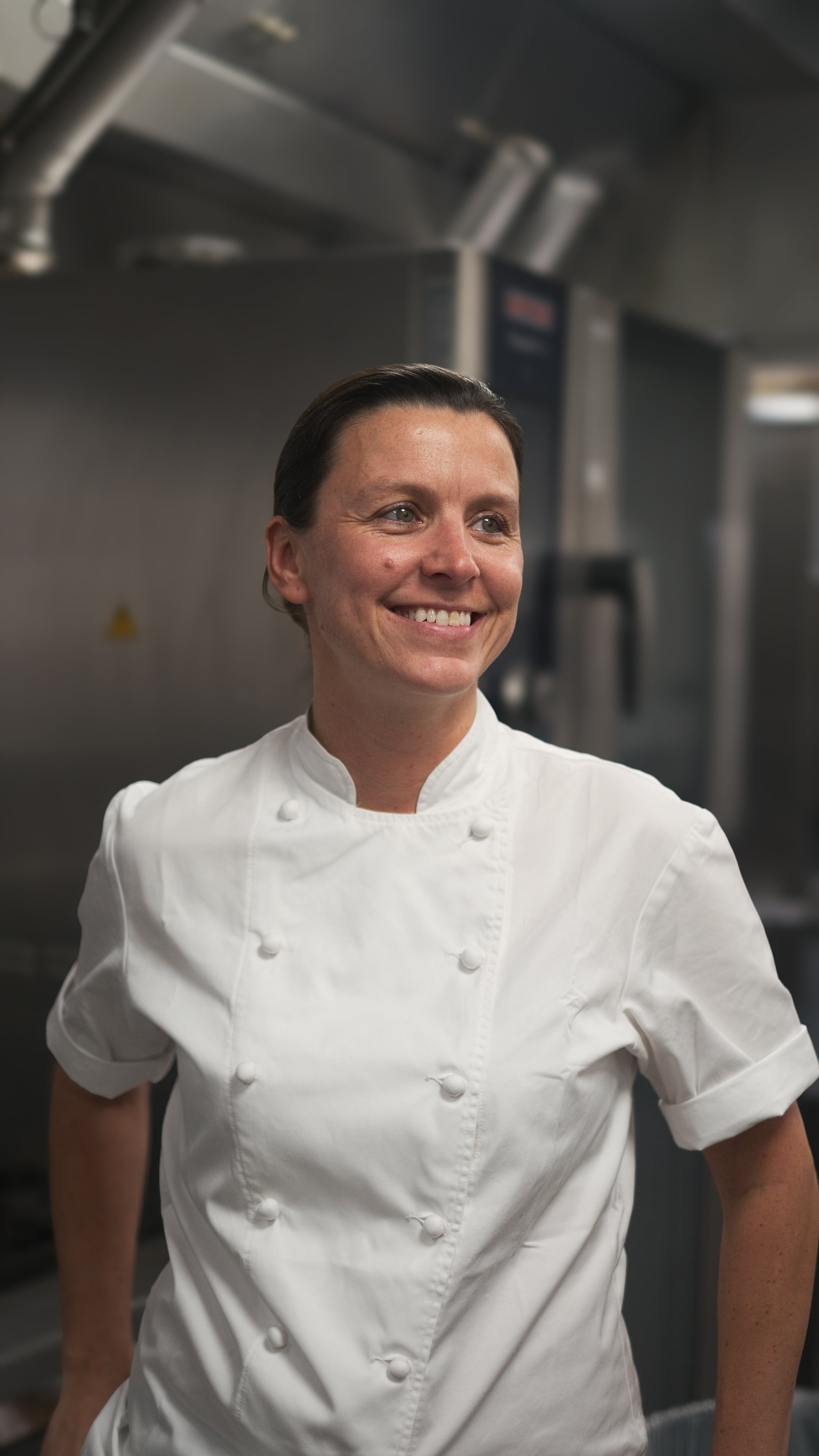 A smiling woman with dark hair pulled back, wearing a white chef's jacket, looking to her left.