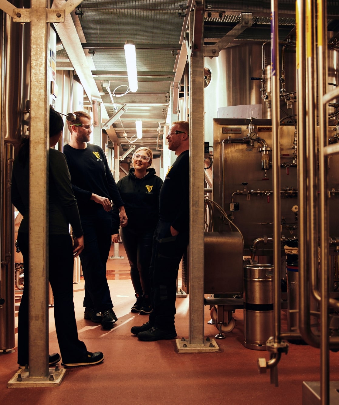 A group of four people, two men and two women, stand chatting in a brewery.
