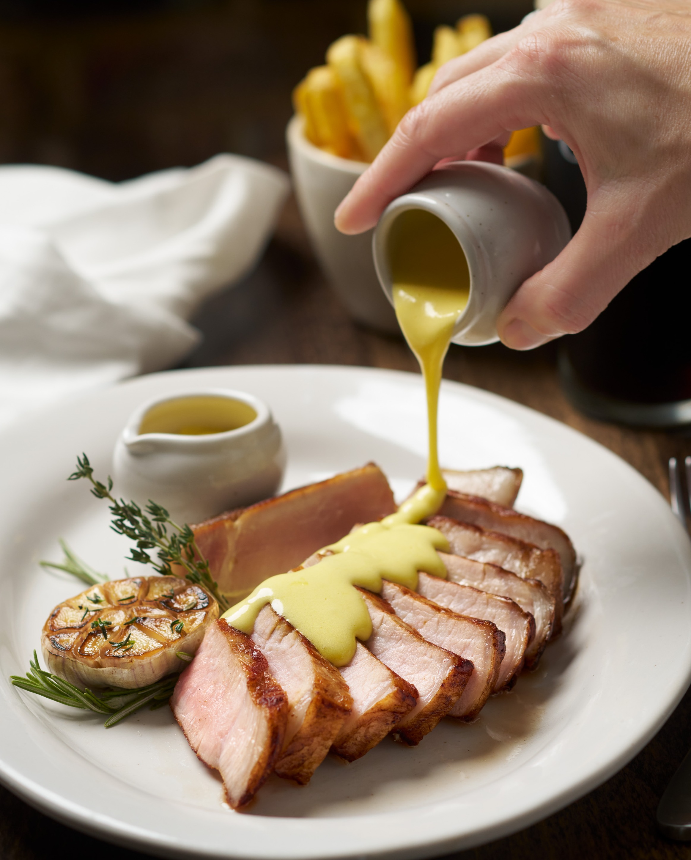 A hand pours yellow sauce over sliced pork chops, garnished with roasted garlic and herbs, on a plate with fries, a dark drink, and cutlery.