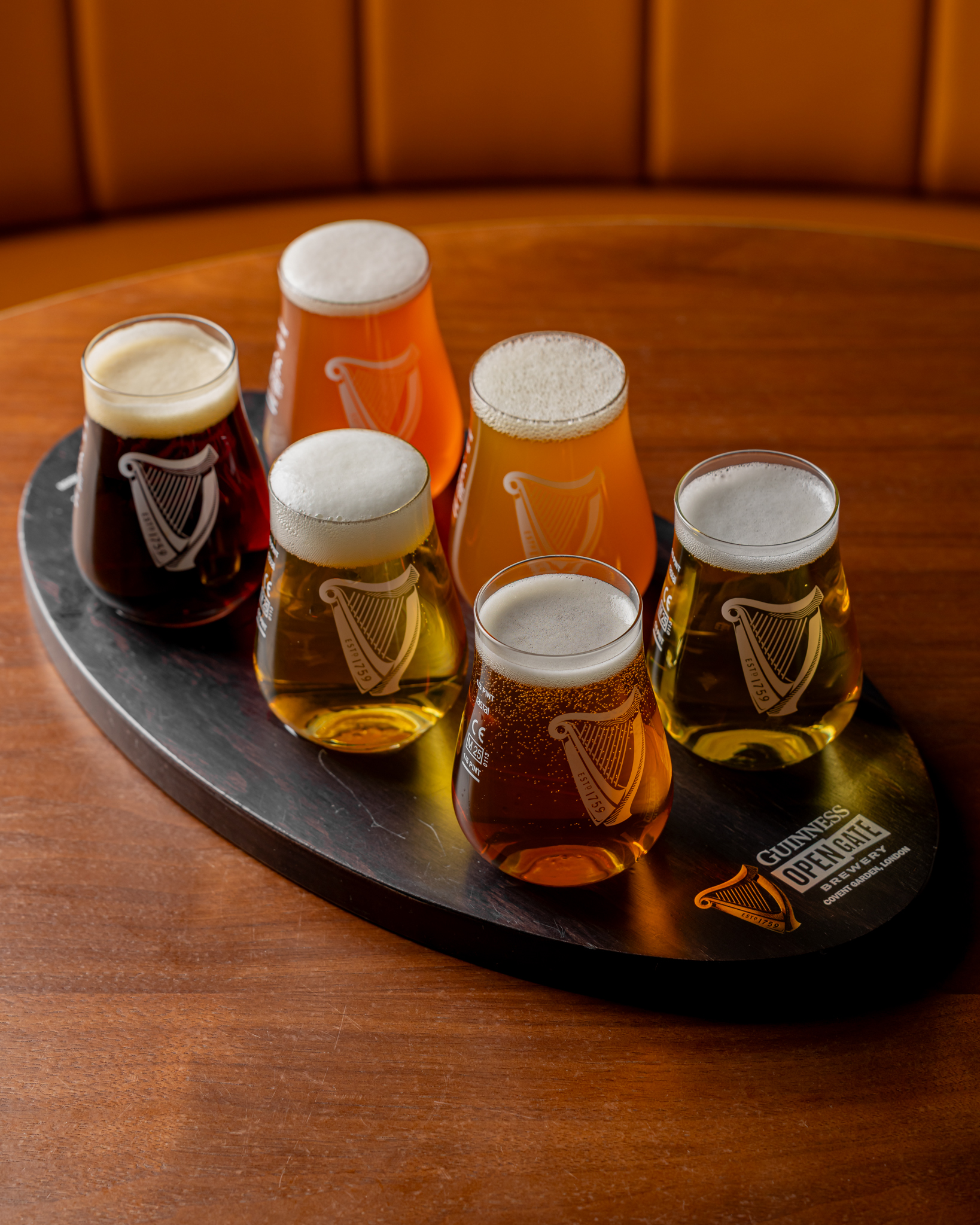 A brown wooden table with a beer flight of 6 beers on a oval black wooden Guinness branded flight