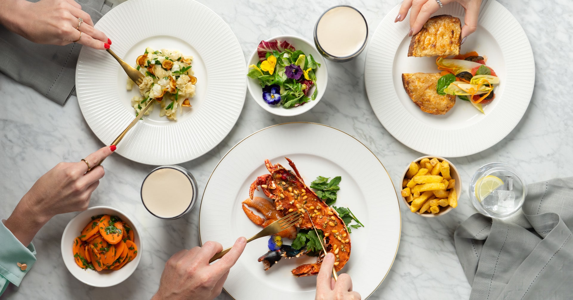 A table laden with various dishes, including lobster, a burger, fries, salads, and drinks, with multiple hands reaching for the food.